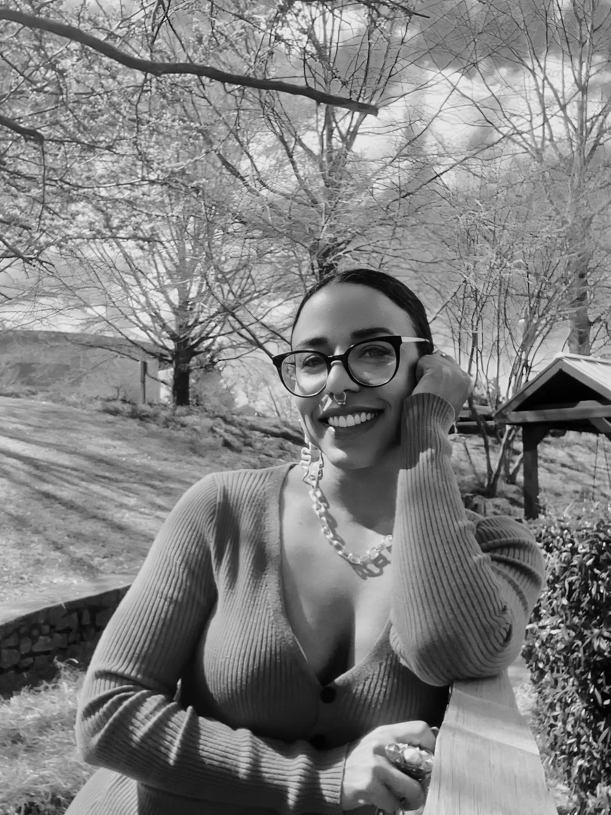 A black and white photo of Tatiana Lima wearing glasses, smiling with her right hand on her cheek.