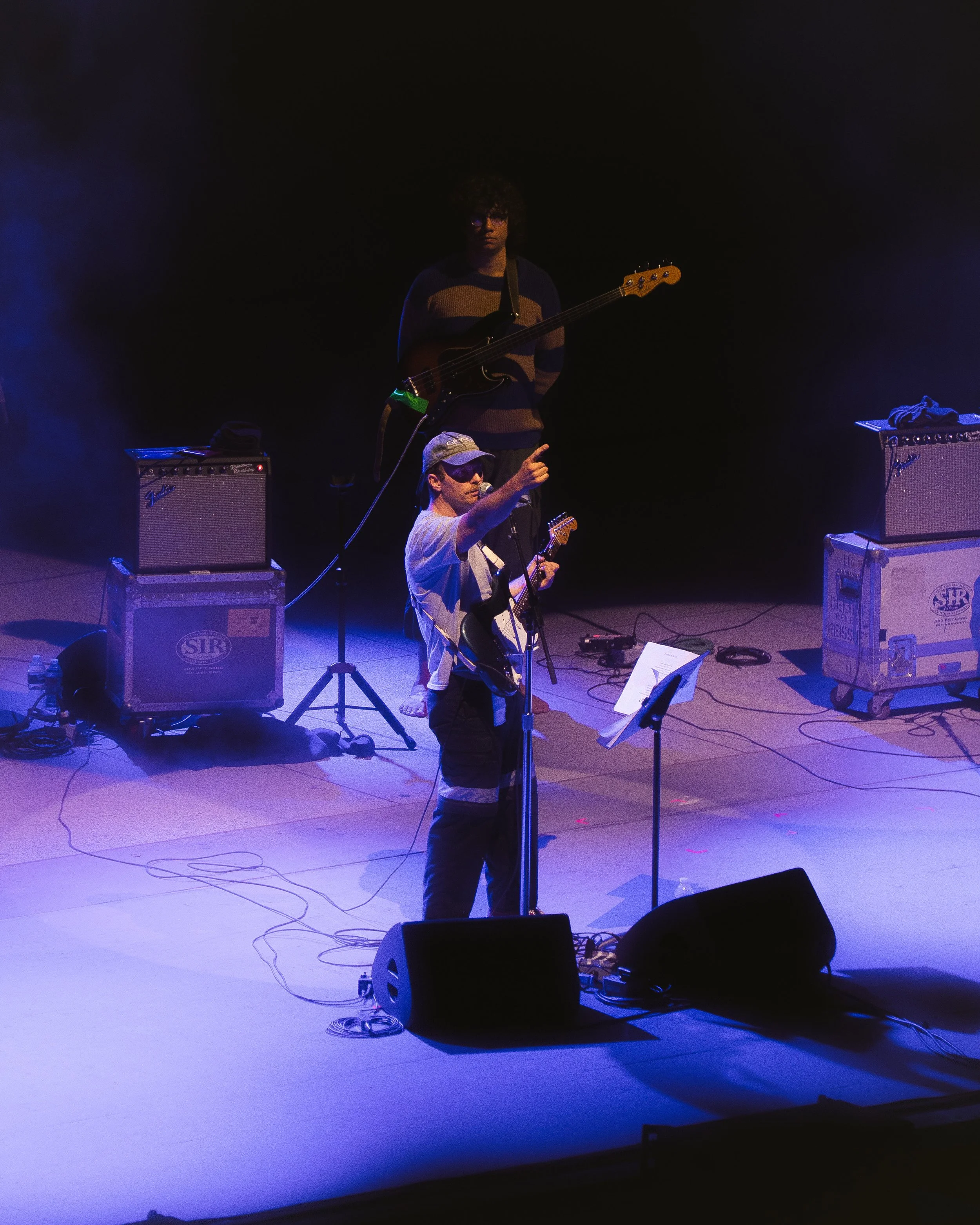 Two musicians perform on stage in a dark setting with purple and blue lighting. One is playing an electric guitar and singing into a microphone, wearing a striped shirt and cap, while the other is standing behind with a bass guitar, wearing a striped