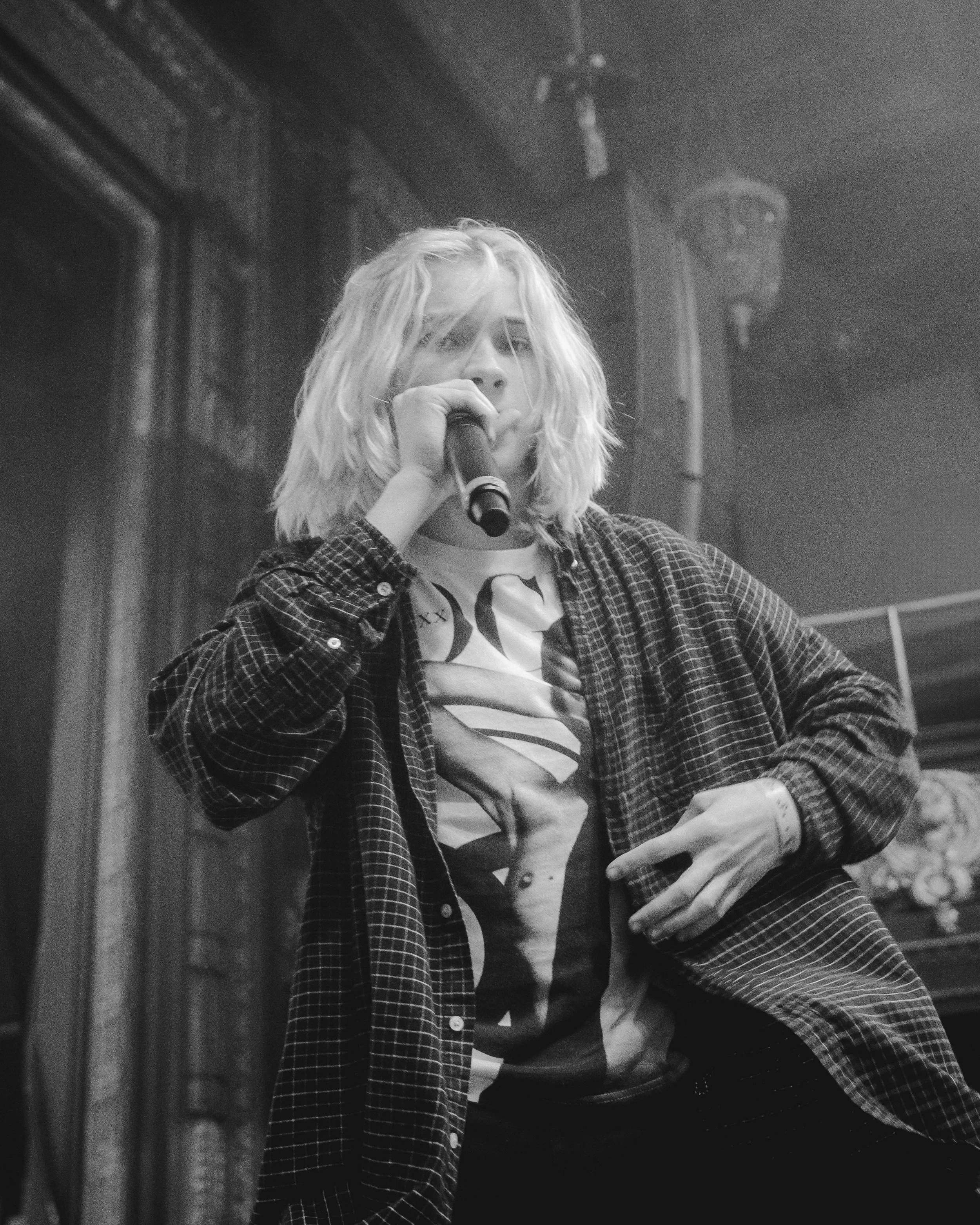 A young woman with shoulder-length blonde hair holds a microphone near her mouth, wearing a checkered shirt over a graphic t-shirt, standing in front of a dark wall with a lamp in the background.