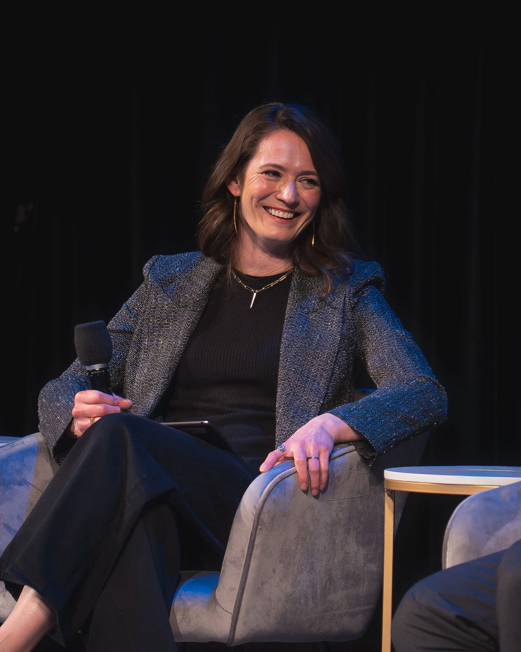 A woman with brown hair, smiling while holding a microphone, sitting at a table during a panel discussion or interview. She is wearing a textured gray blazer over a black top, gold jewelry, and has a dark background behind her.