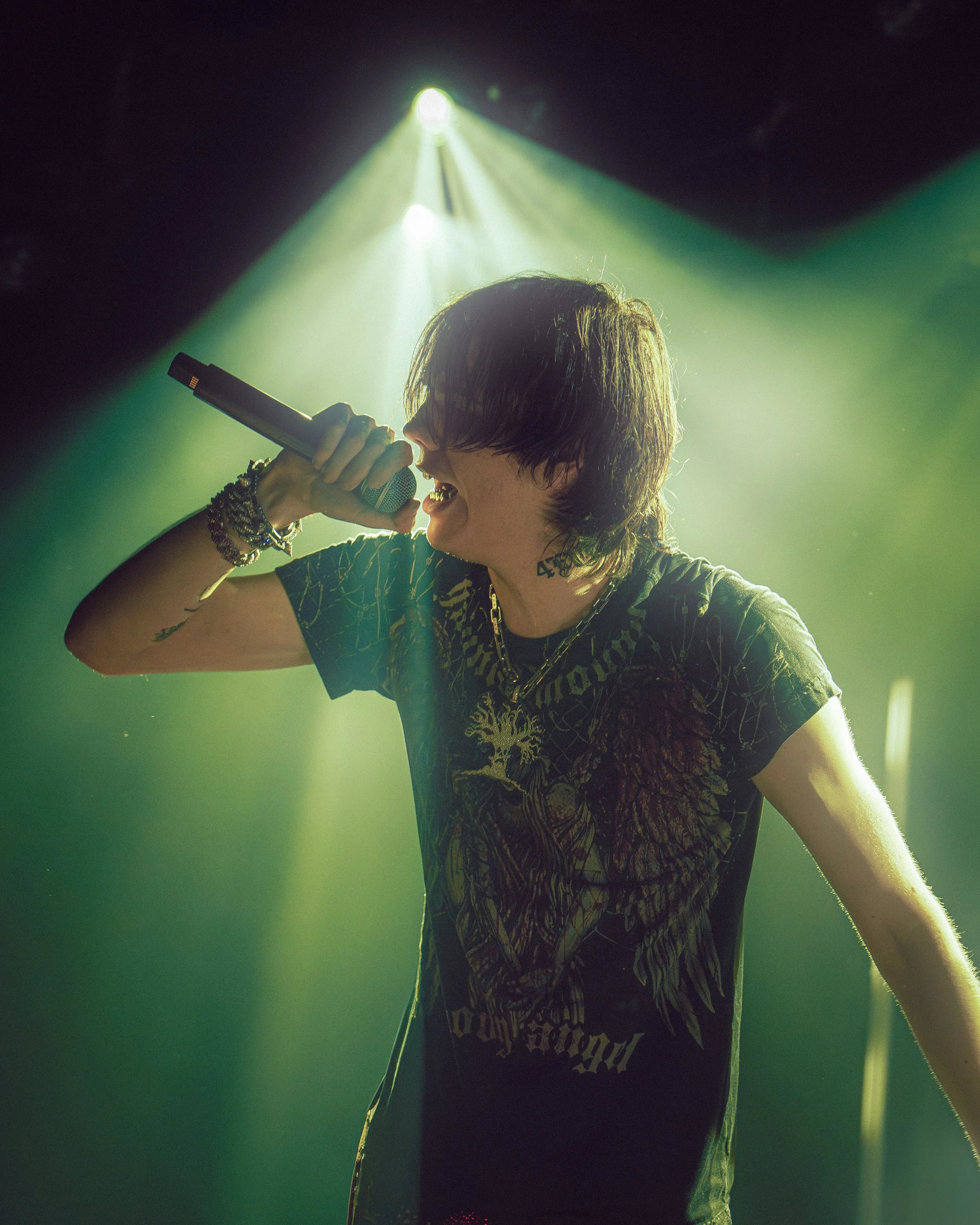 A person singing into a microphone on stage, illuminated by green and yellow stage lights, wearing a dark graphic T-shirt, glasses, and jewelry.