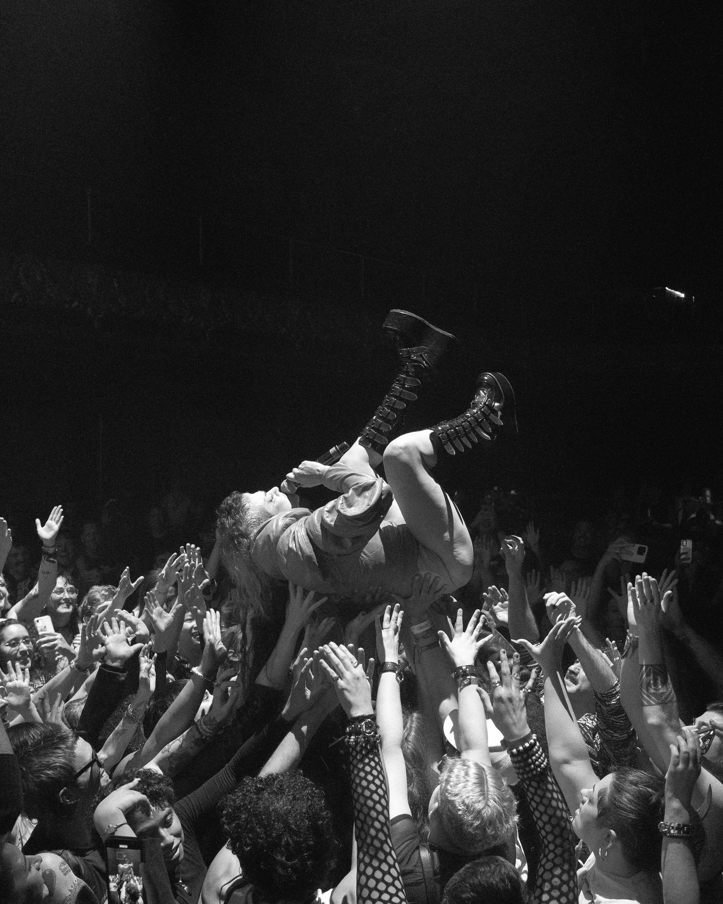 A large crowd of people gathered at an outdoor event at night, with many reaching out their hands and some people lifting an individual in the center above the crowd. The person being lifted appears to be male, wearing a jacket, and has short hair. T