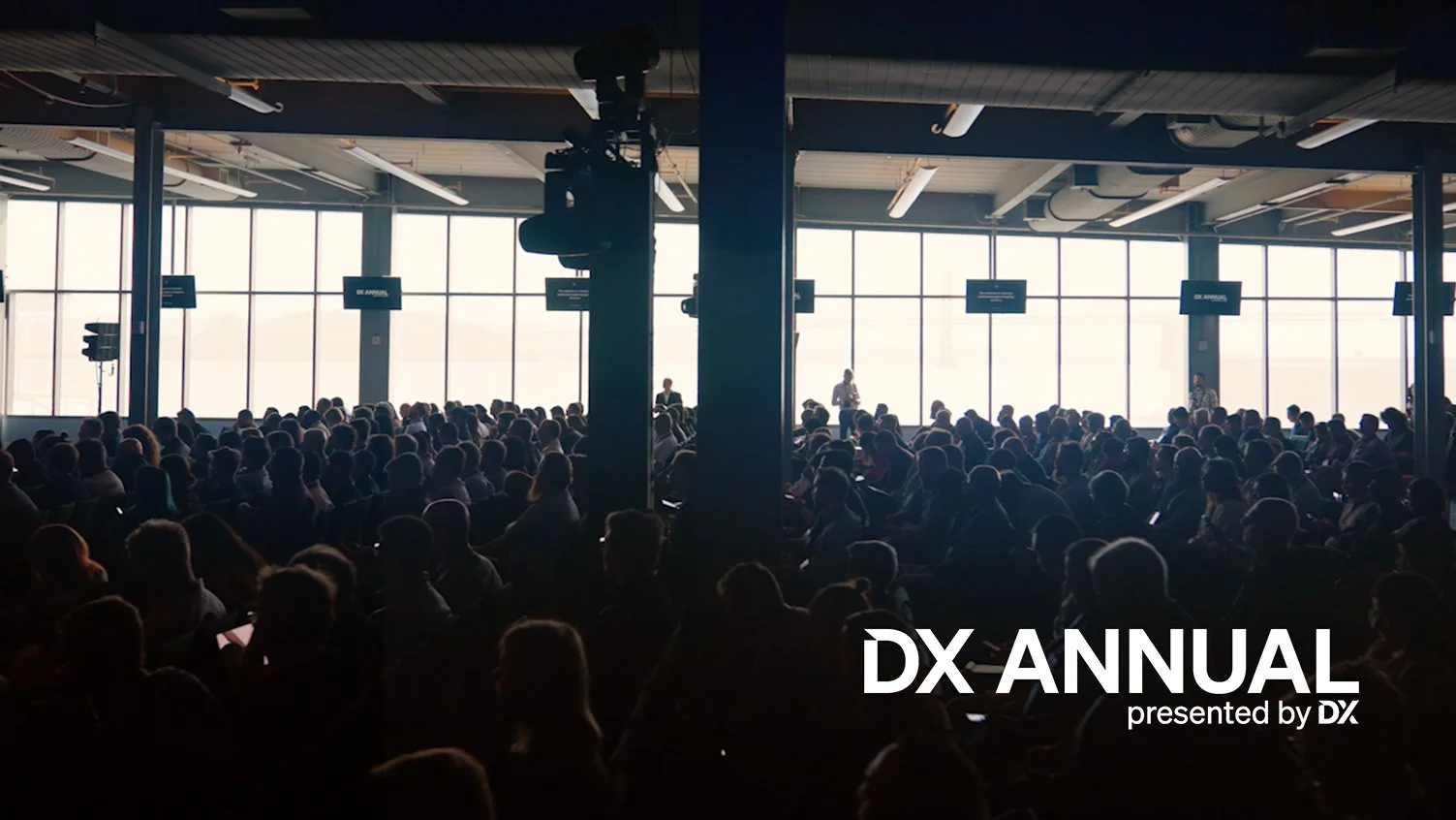 Large audience seated in an indoor conference hall with large windows, during the DX Annual conference presented by DX.