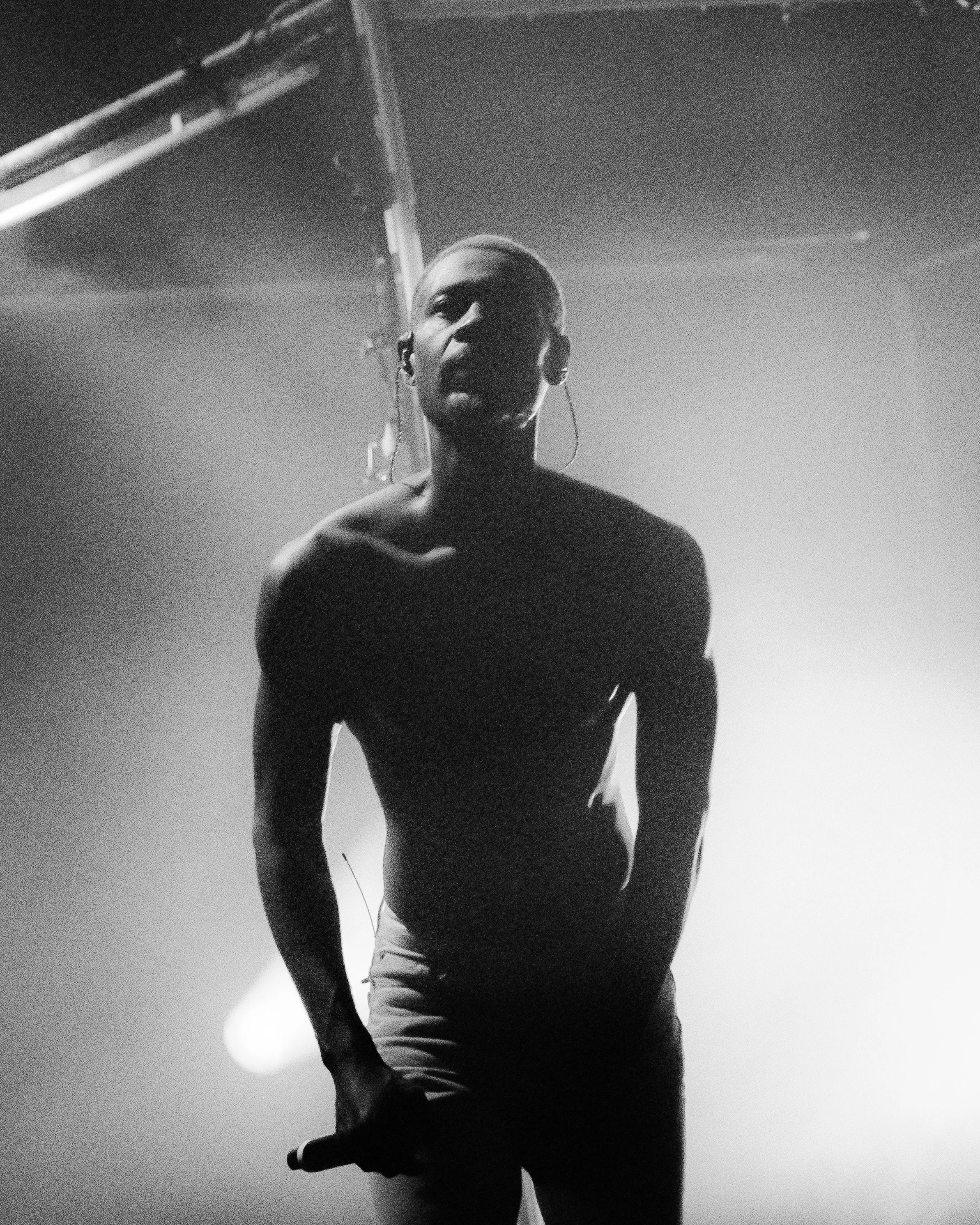 A black and white photo of a shirtless man with earrings, standing in a dimly lit room with a ladder in the background, facing forward.