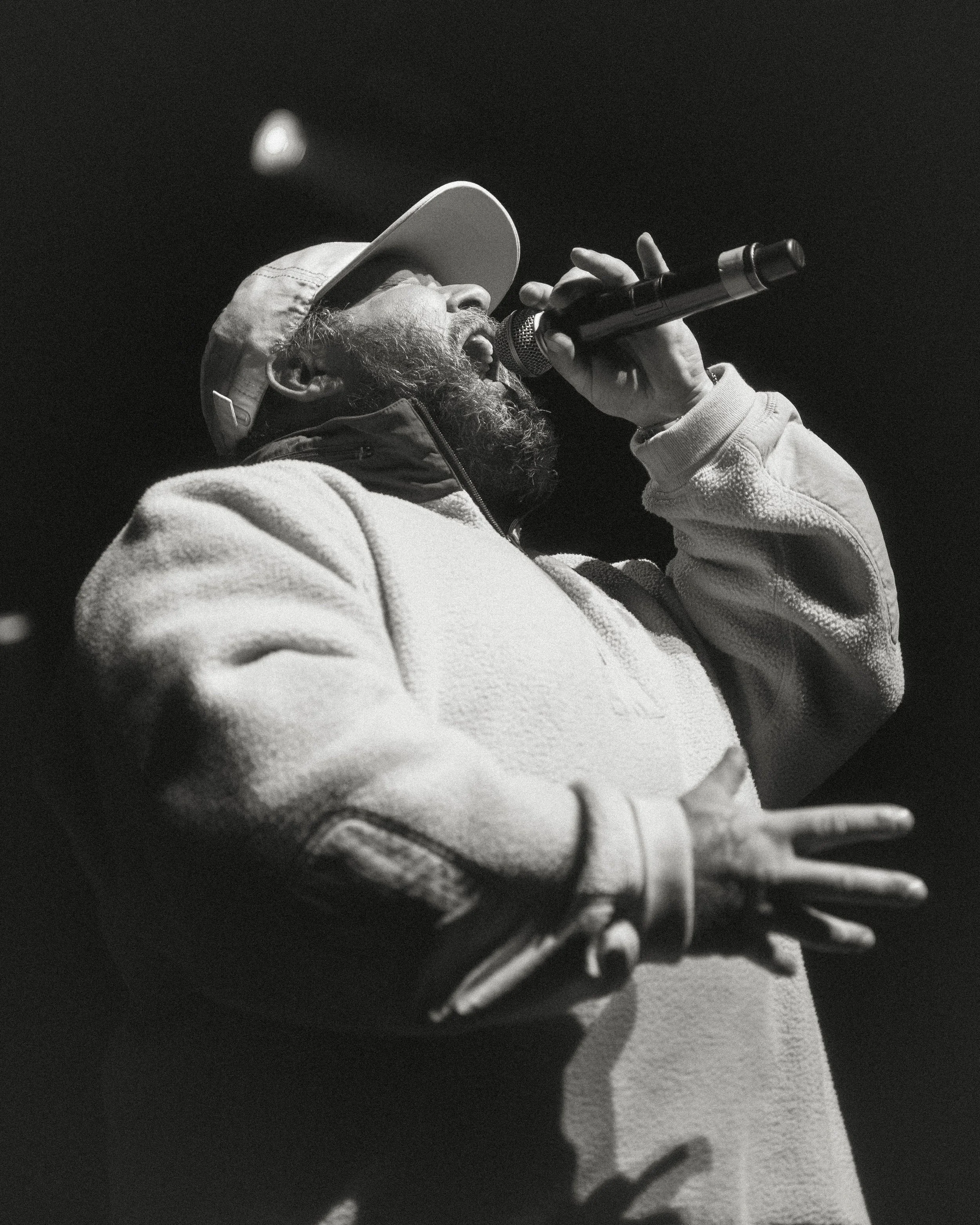 Black and white photograph of a man with a beard wearing a light-colored baseball cap and hoodie, singing into a microphone with his eyes closed against a dark background.