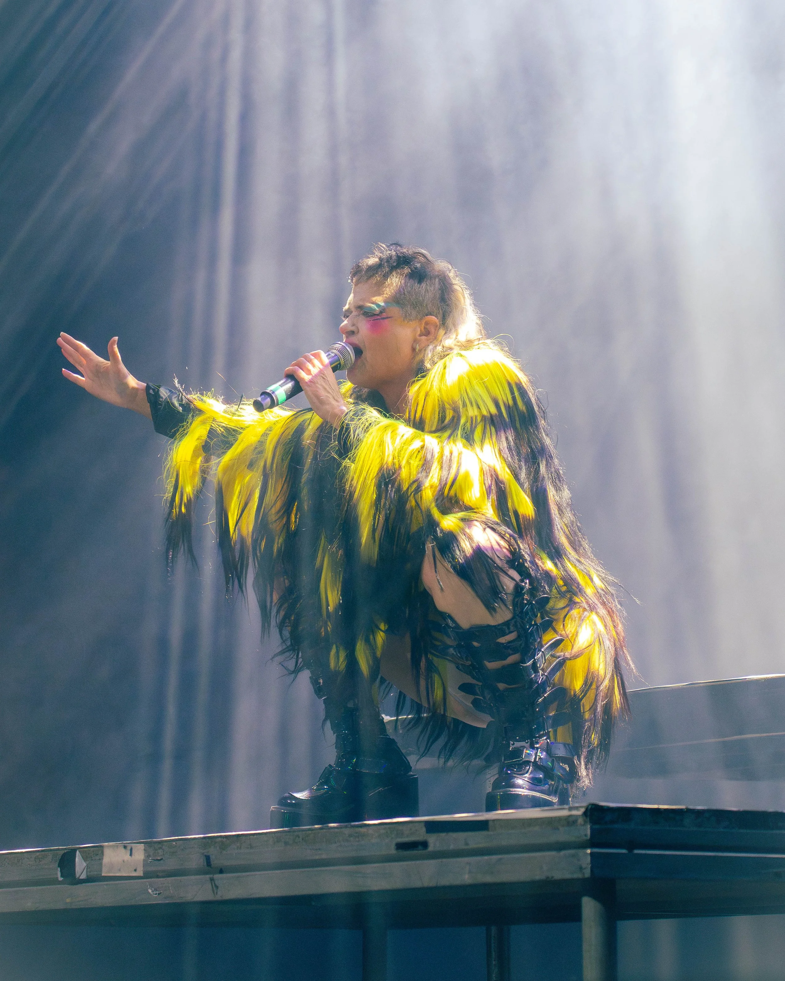 A performer dressed in a yellow feathered outfit singing into a microphone on stage, with dramatic lighting creating rays of light in the background.
