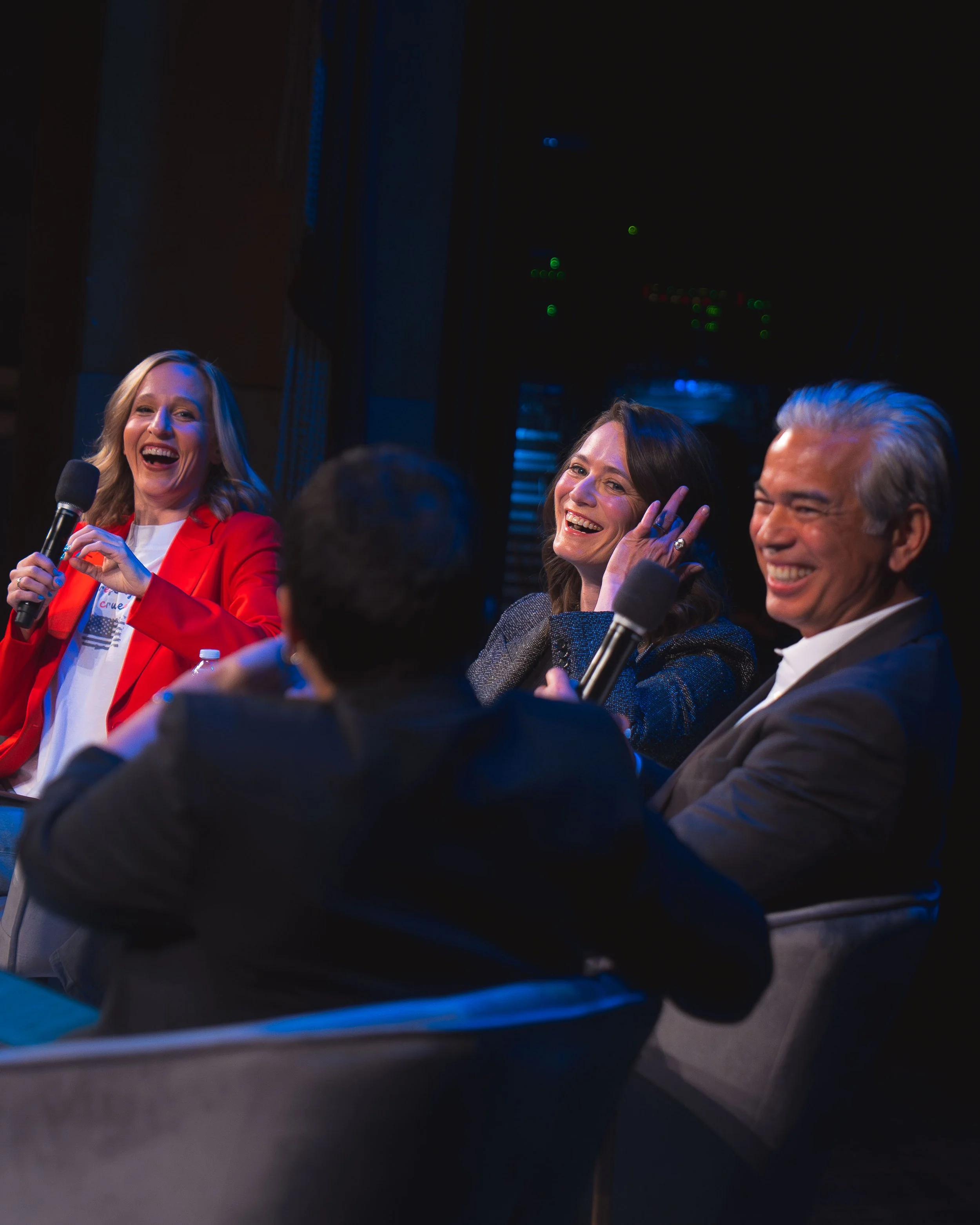 Three smiling people sitting together, one woman holding a microphone, in a dark room with dim lighting.