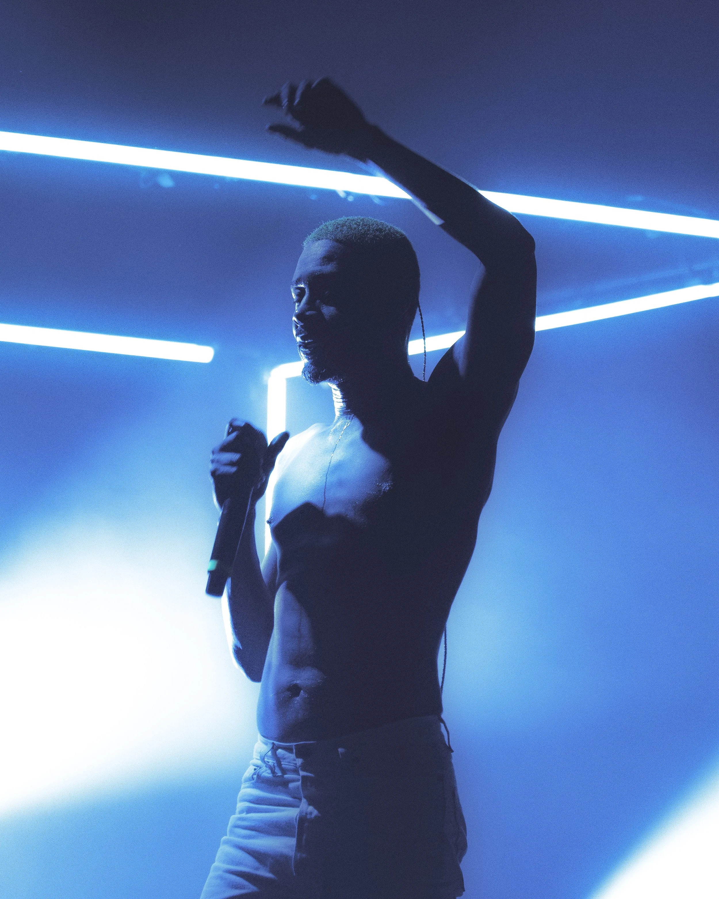 A person with short hair and a beard, wearing a sleeveless shirt and pants, appears to be dancing or performing in a dark environment with blue neon lights.