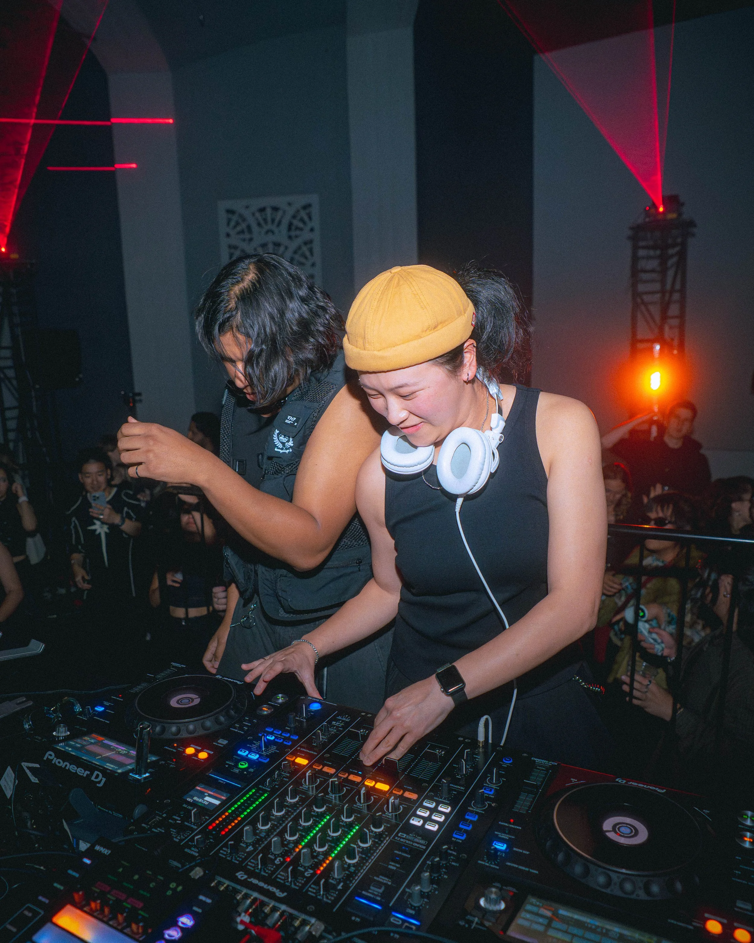 Two DJ performers, one wearing a yellow beanie and headphones around her neck, are actively mixing music on a DJ controller with colorful LED lights. They are surrounded by a crowd at a music event with stage lights and laser beams in the background.