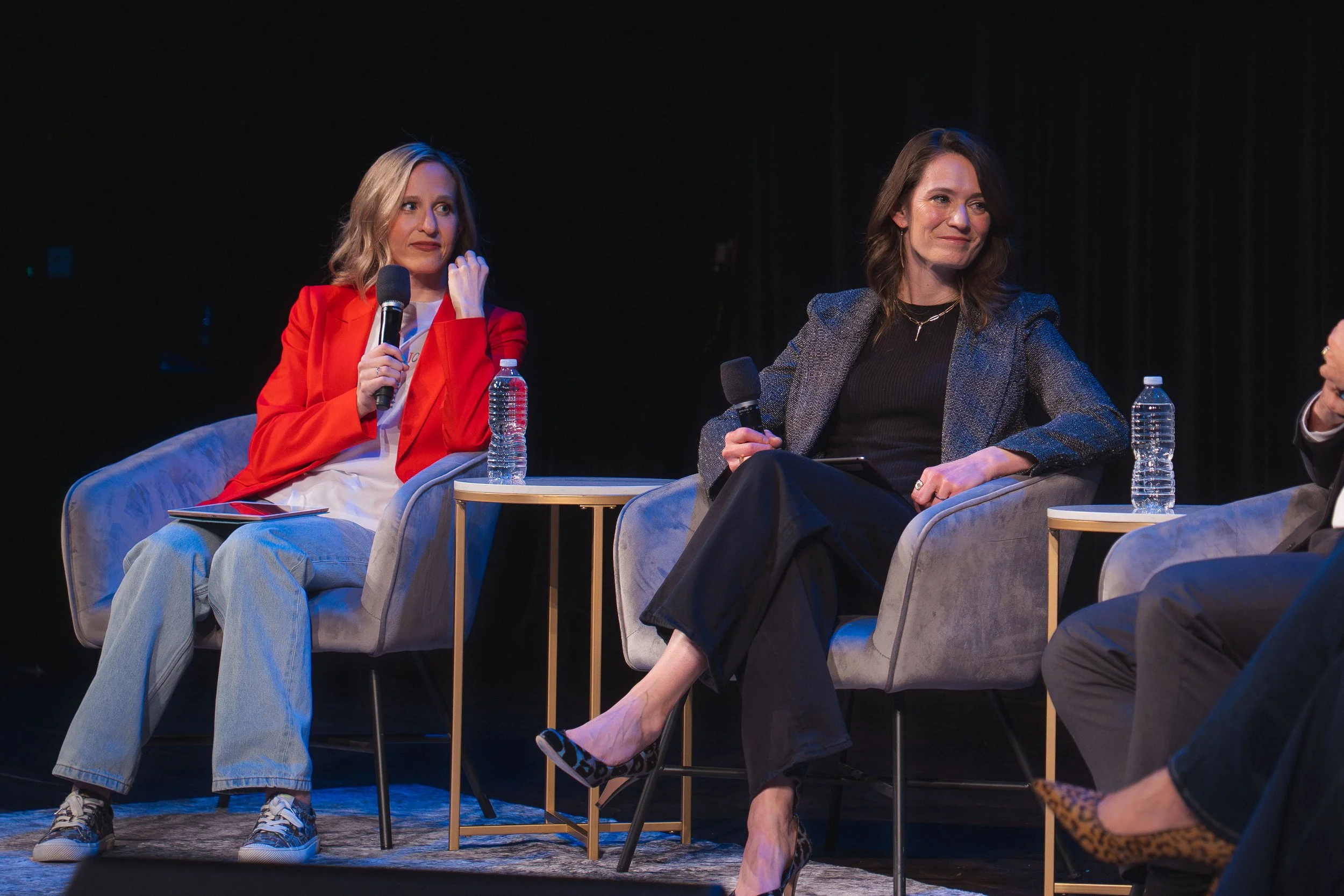 Two women seated in armchairs on stage during a panel discussion, each holding a microphone and water bottle.