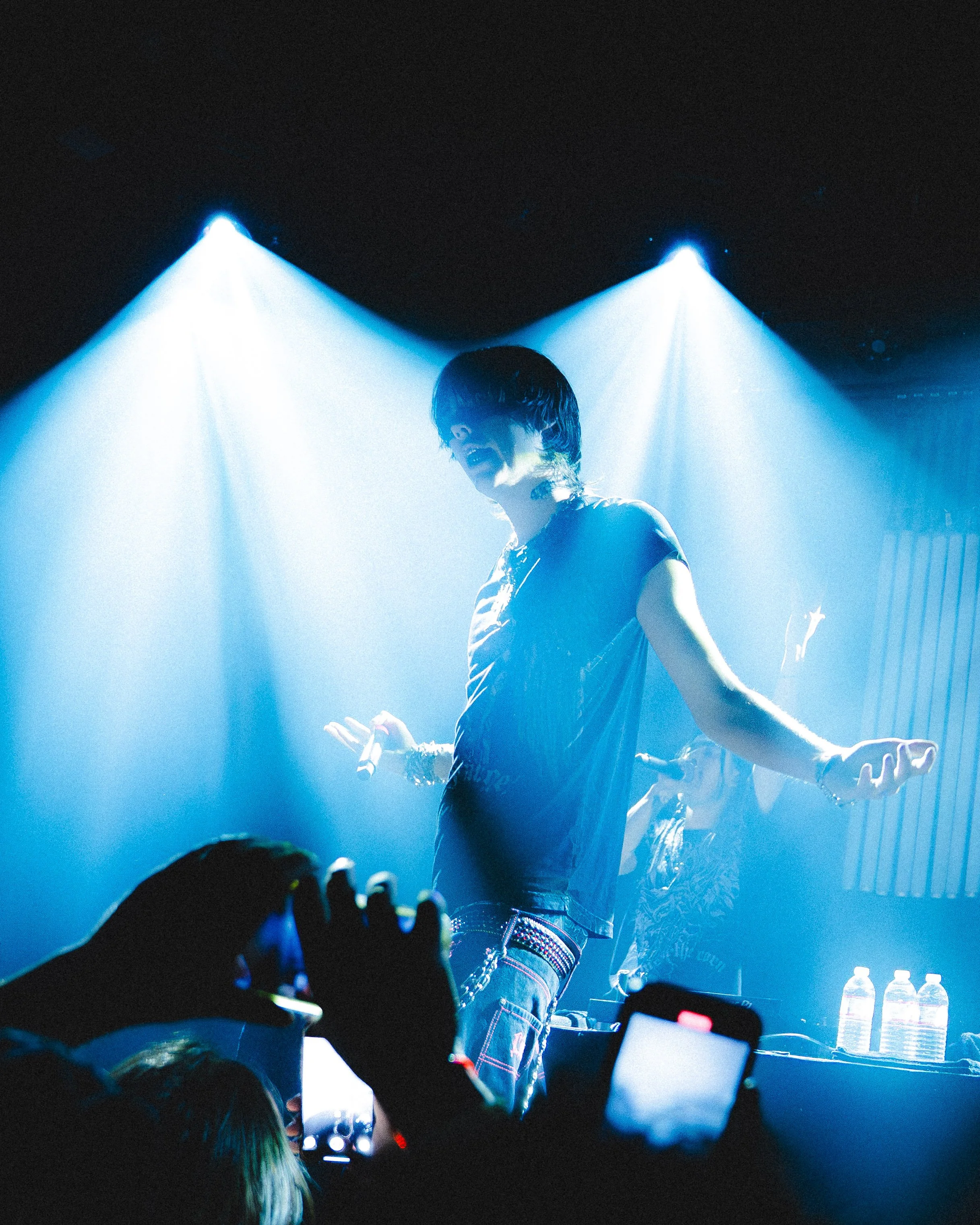 A performer with dark hair in a black shirt on stage illuminated by blue spotlights, seen from an audience member's perspective with concertgoers holding up phones.