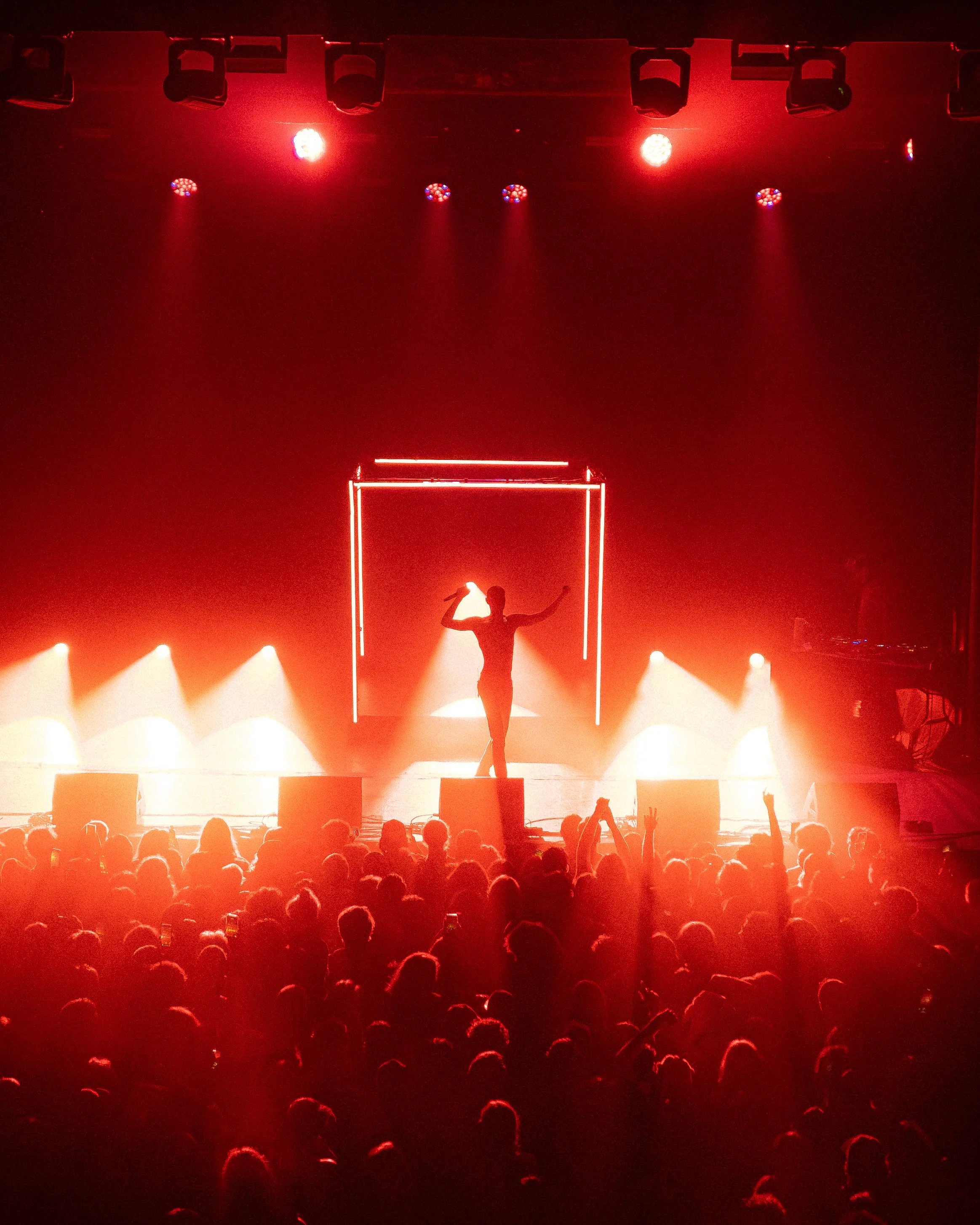 Stage with red lighting, a performer in the center striking a pose, framed by a rectangular neon outline, and a crowd watching in the dark.
