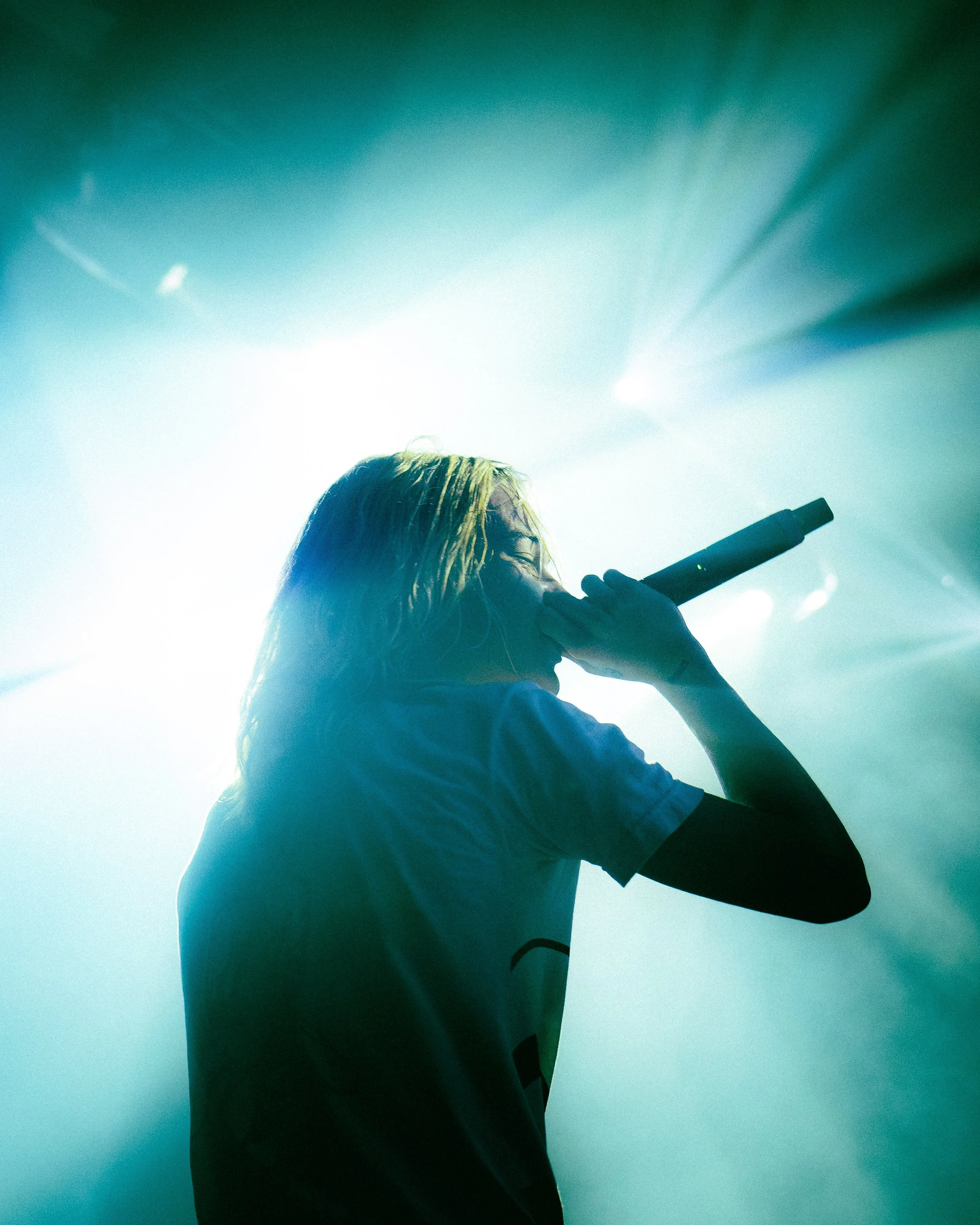 A silhouette of a person with long hair looking through a microscope against a bright, illuminated background with teal and white hues.