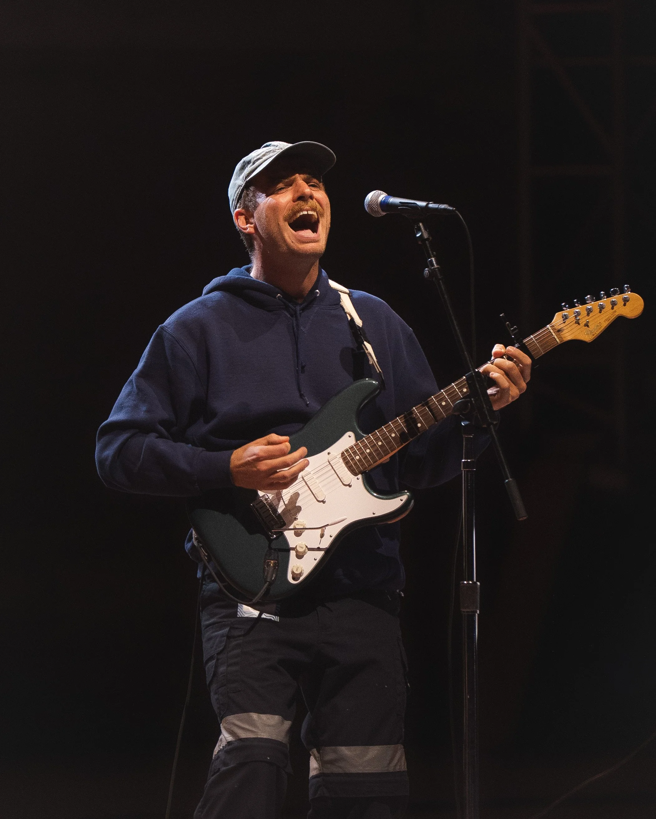 Man wearing a gray cap and black hoodie singing passionately while playing an electric guitar in front of a microphone on a dark stage.