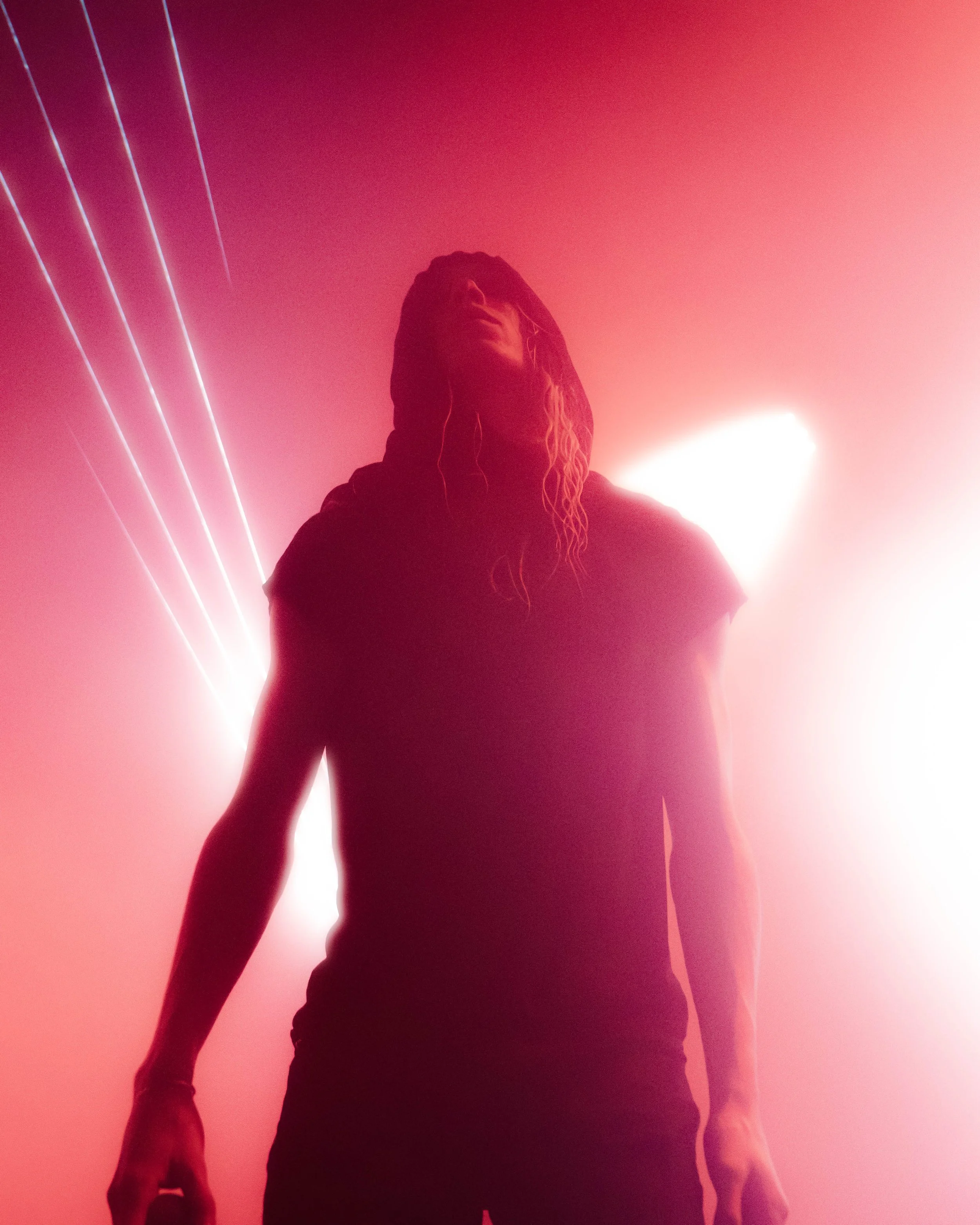 A person with dreadlocks wearing a hoodie stands against a vibrant pink and red background with bright lights, creating a silhouette effect.