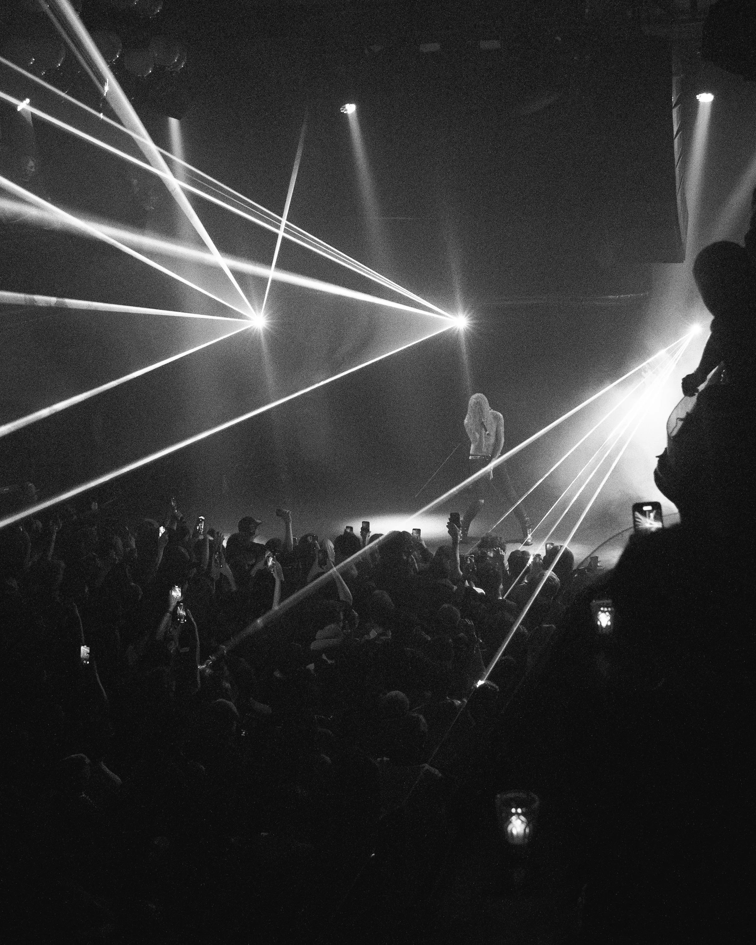 A concert scene with multiple bright laser lights illuminating the stage and audience. Silhouettes of people are visible, some holding phones to capture the moment. The stage features a performer, possibly a singer or musician, with some details obsc
