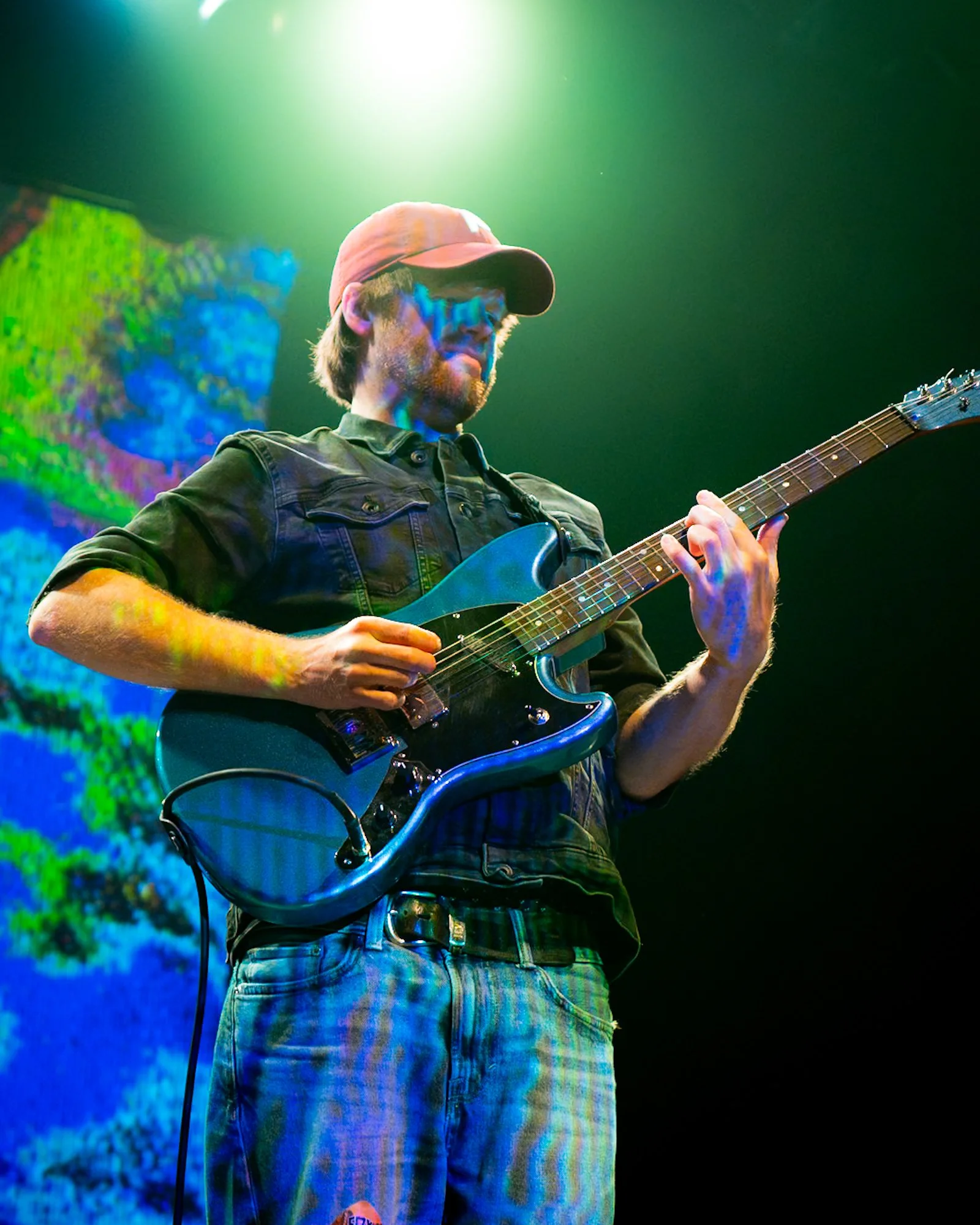 A man wearing a brown hat and sunglasses playing an electric guitar on stage, with colorful stage lights and a digital screen behind him.