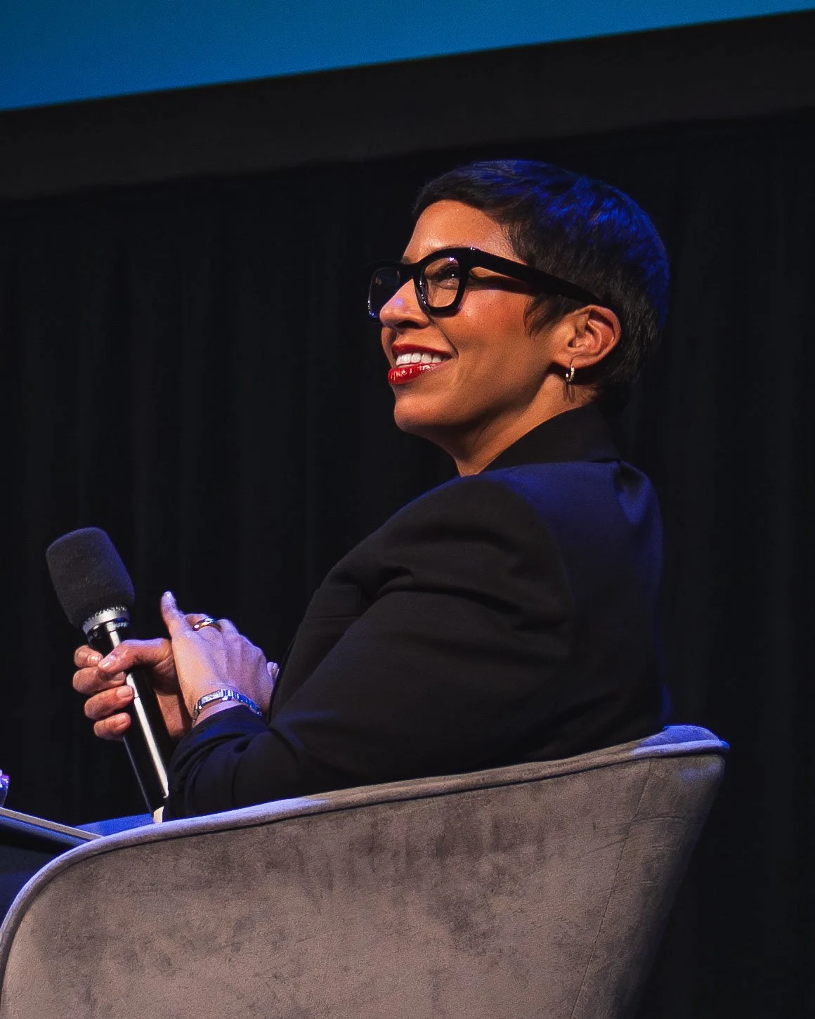 A person with short dark hair, glasses, and earrings, smiling and holding a microphone, seated on a stage against a dark curtain background.