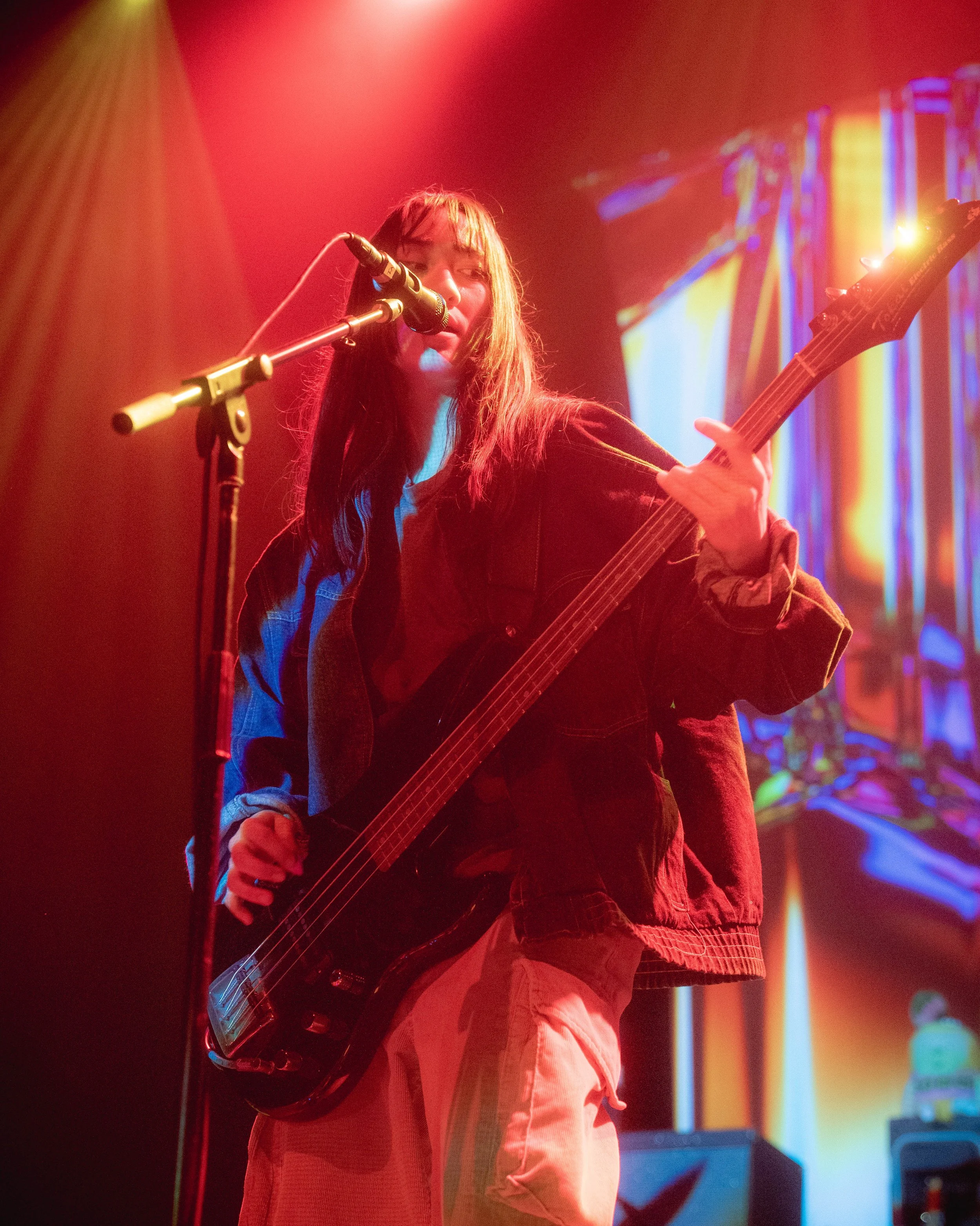 A young woman with long hair is playing an electric guitar and singing into a microphone on stage, with colorful stage lights and a digital screen in the background.