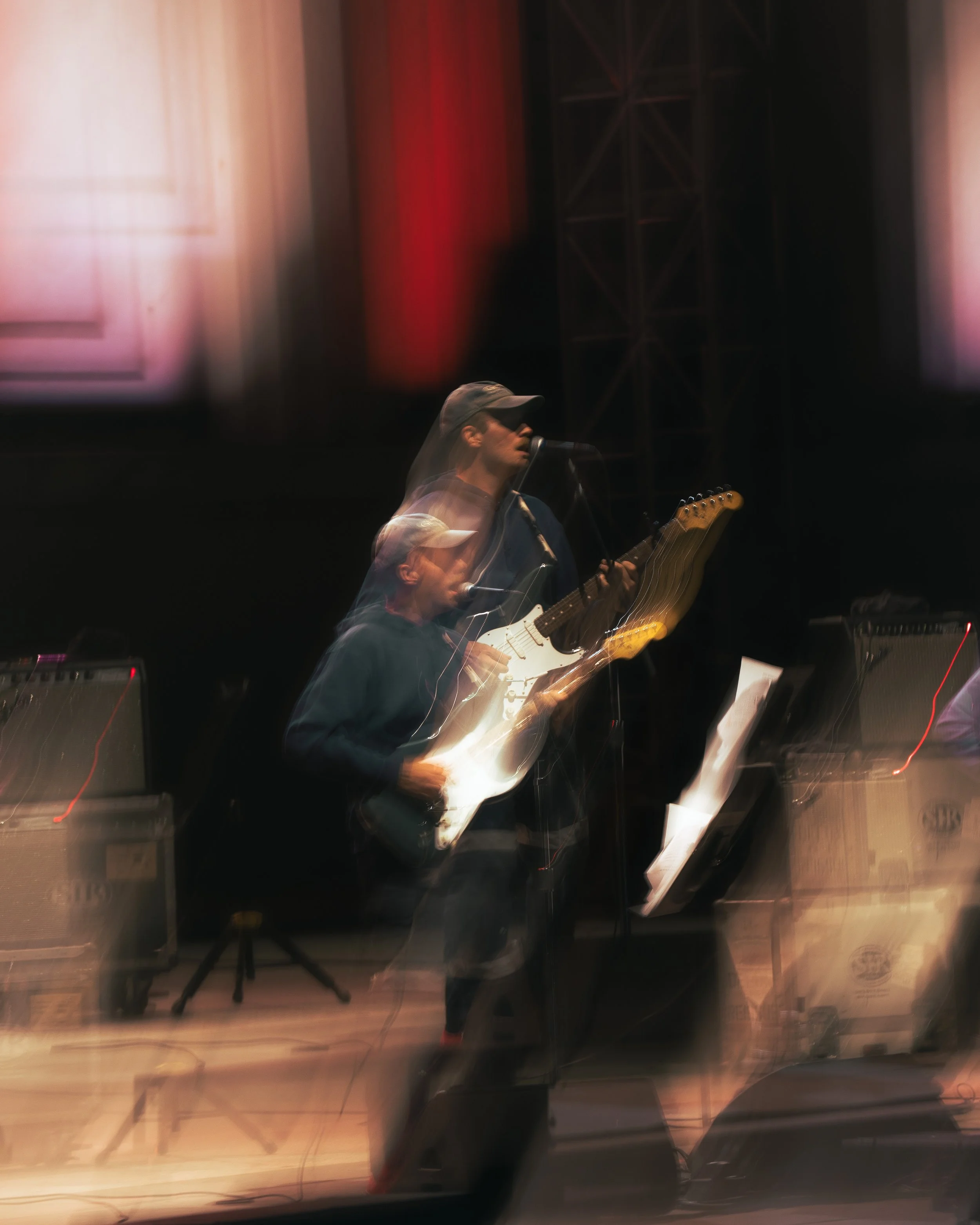 A musician playing an electric guitar on stage, with a blurred effect and colorful lighting, in front of a music stand and amplifiers.
