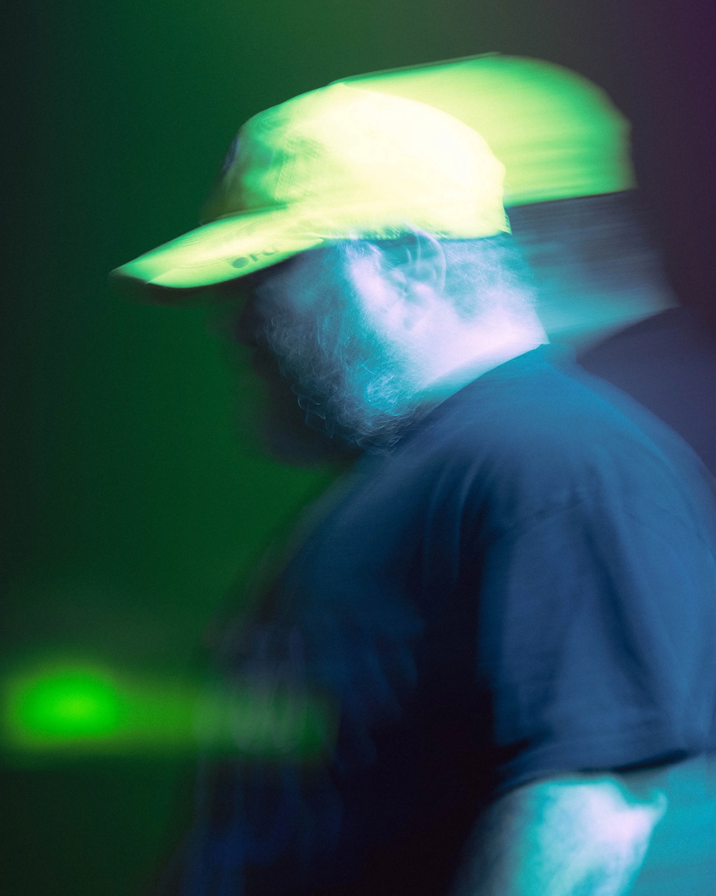 A person wearing a neon yellow cap and black shirt, illuminated by colorful green and blue lights, with a blurry background.