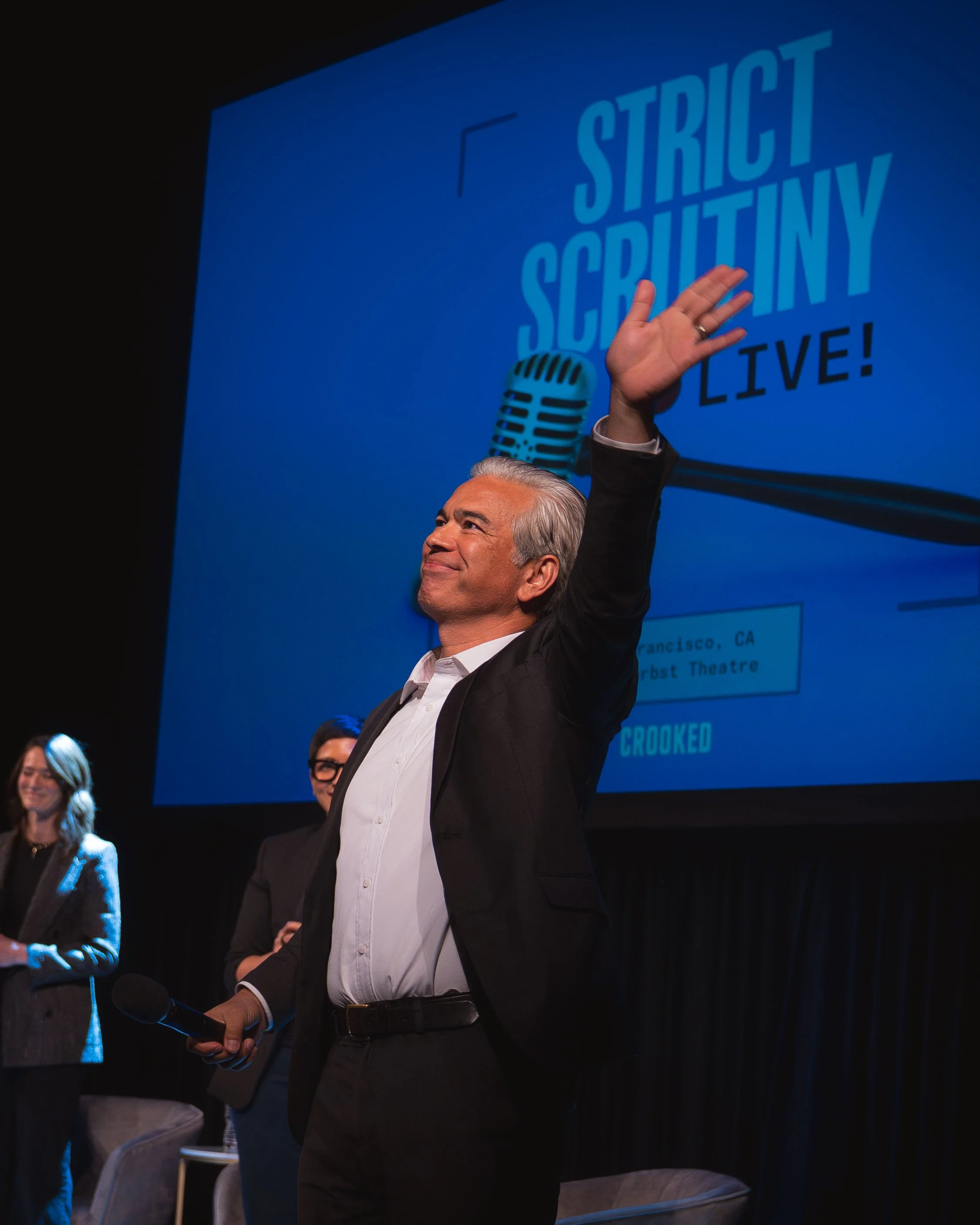 A man in a suit holding a microphone and raising his hand during a presentation with a large blue screen behind him. The screen displays the words 'Strict Scrutiny Live!' and a microphone icon.