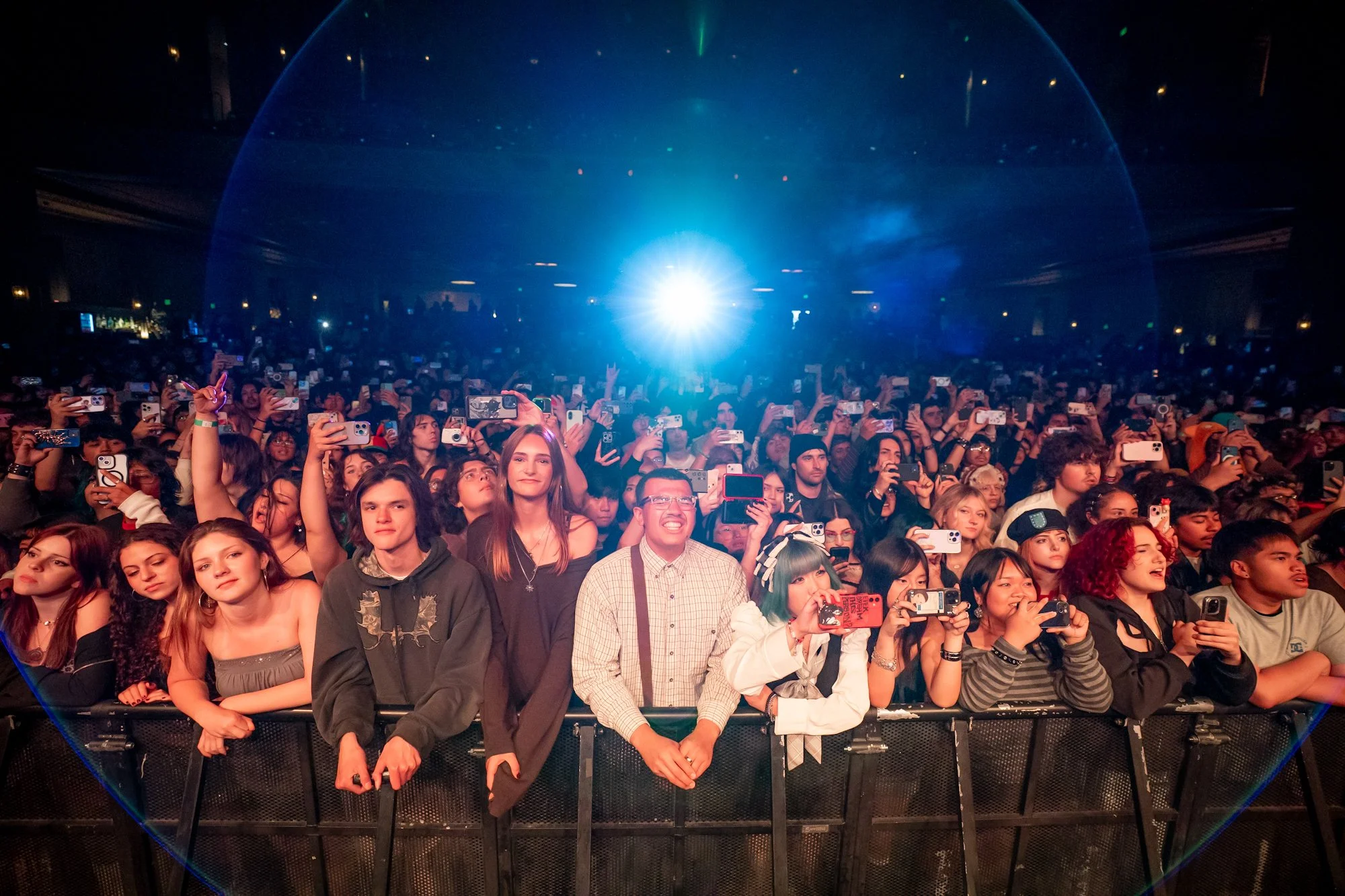Crowd of people at a concert or event, many holding up smartphones, with a bright light on stage creating a blue glow and seeing the audience through a circular frame.