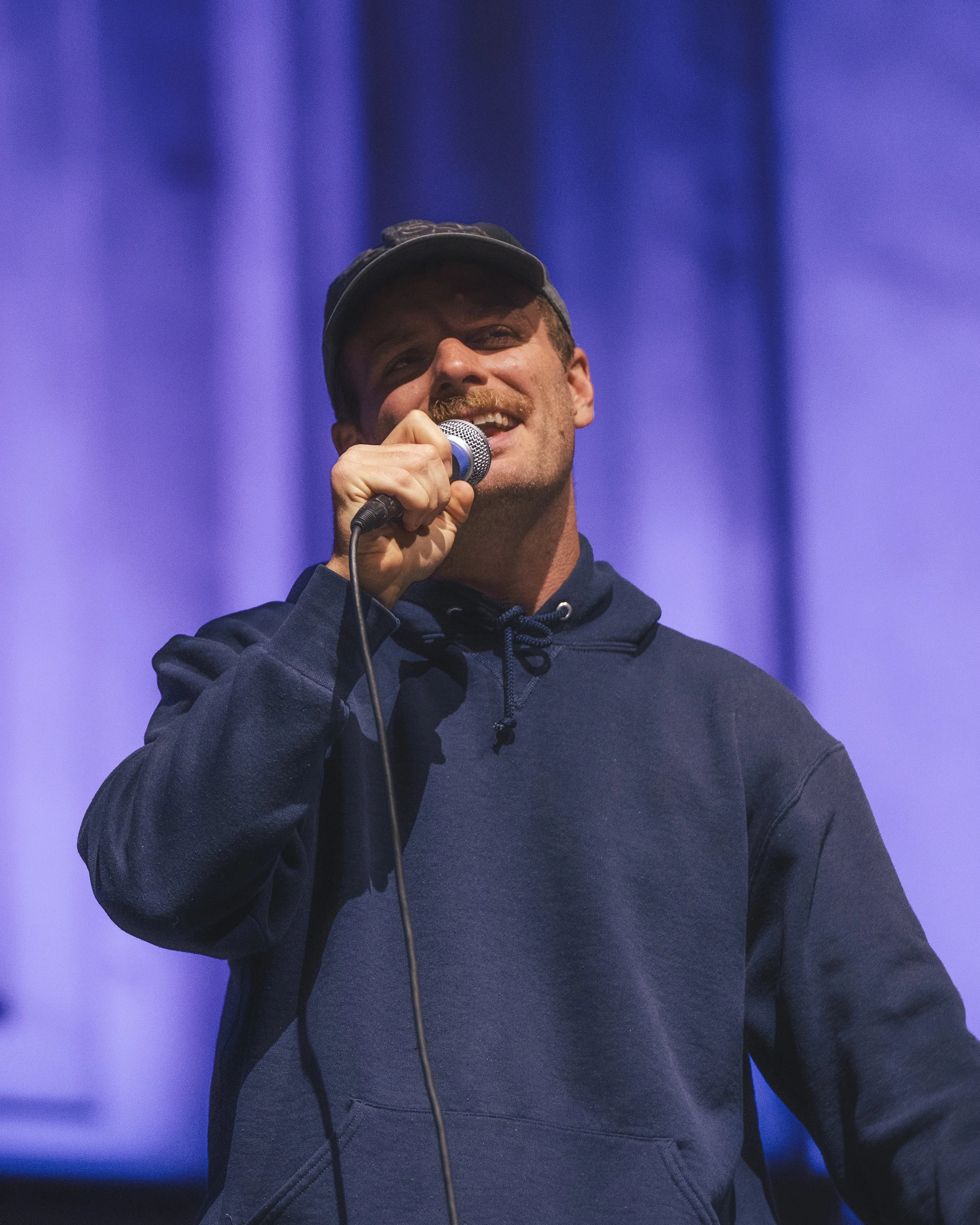 A man wearing a dark hoodie and cap holding a microphone on a stage with a purple background.