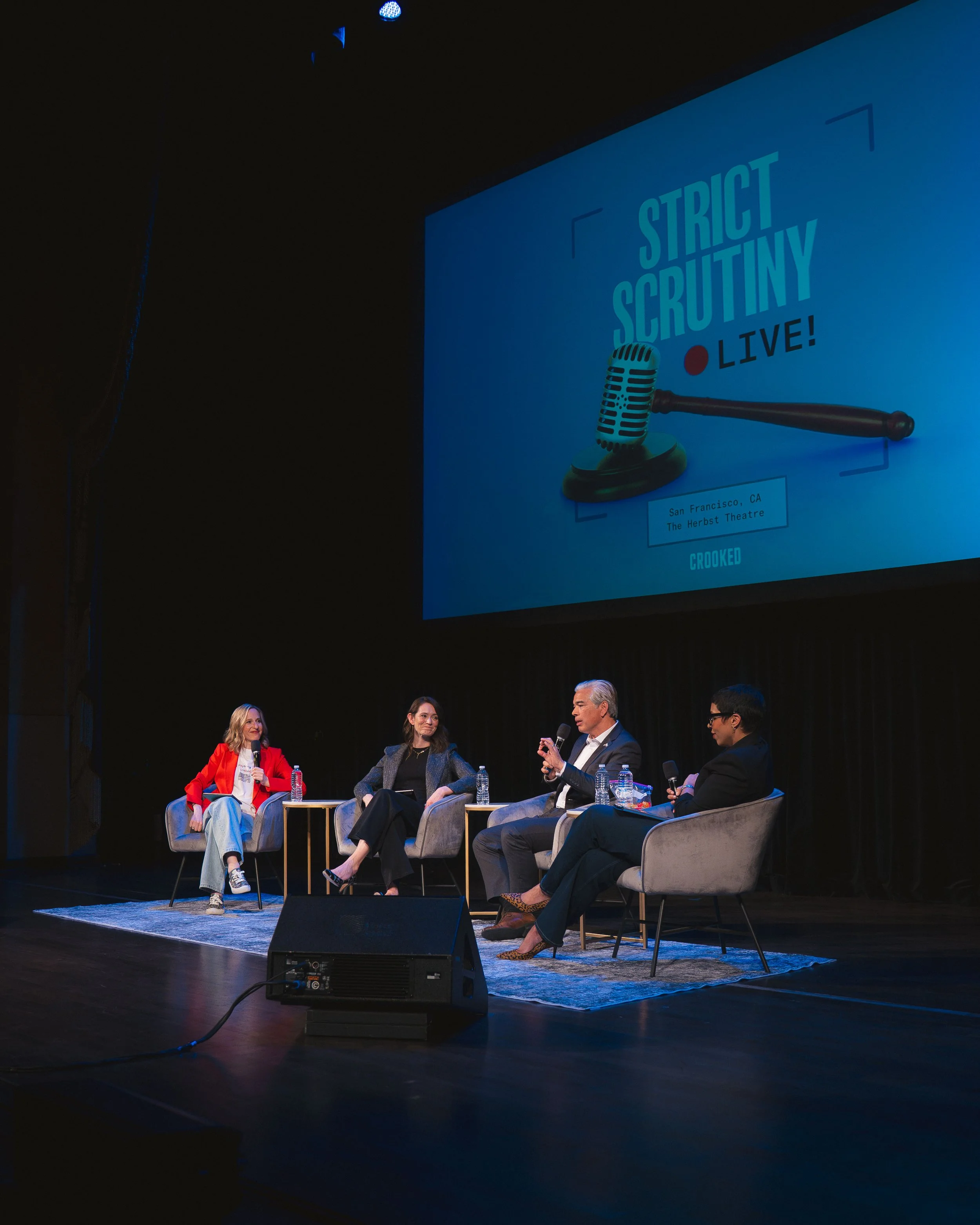 A panel of five people seated on stage during a live discussion or interview, with a large screen behind them displaying the text 'Strict Scrutiny Live!' and a microphone graphic. The stage is set in the Herbst Theatre in San Francisco, CA.