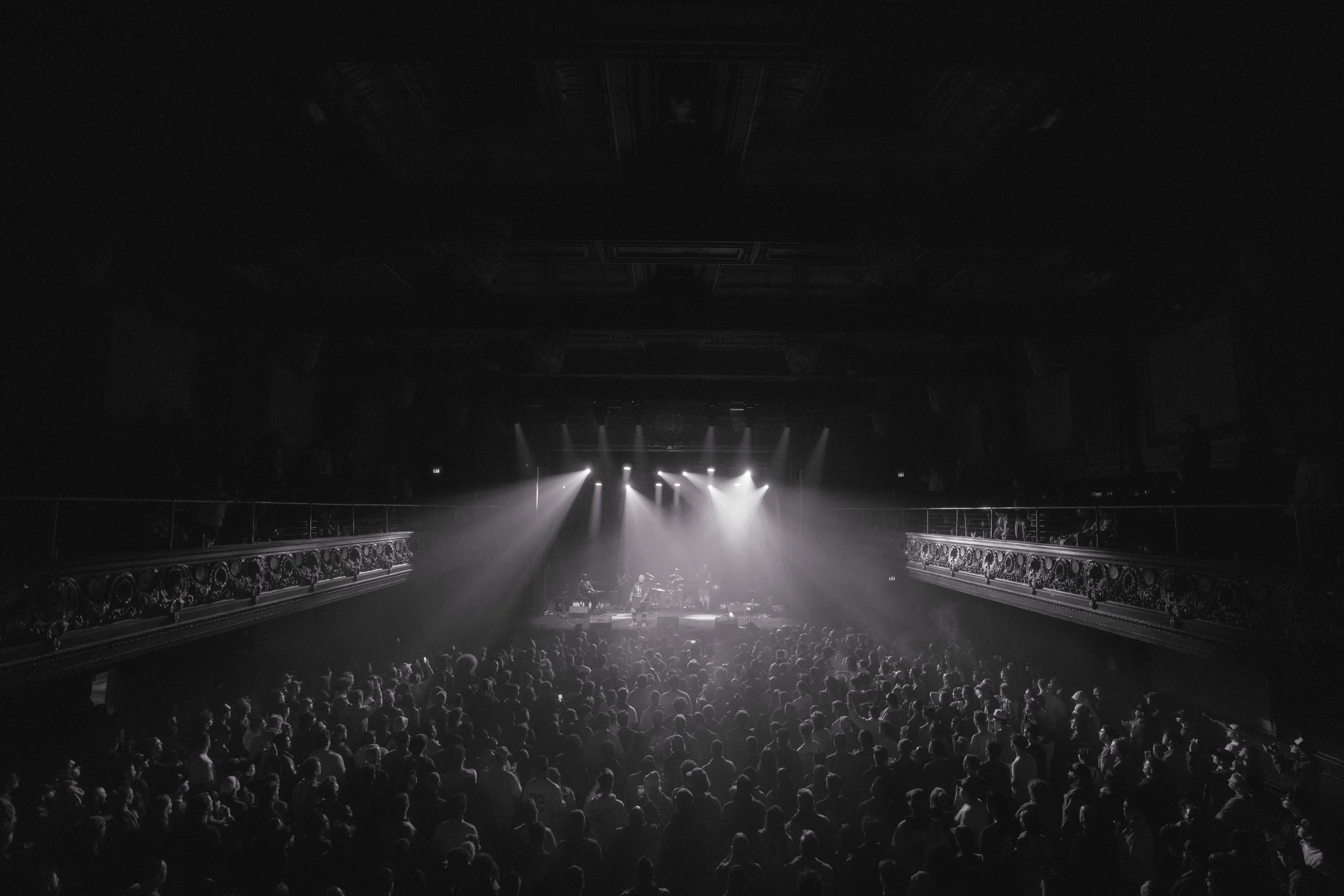 Concert hall with performers on stage and audience seated in front, illuminated by stage lights.