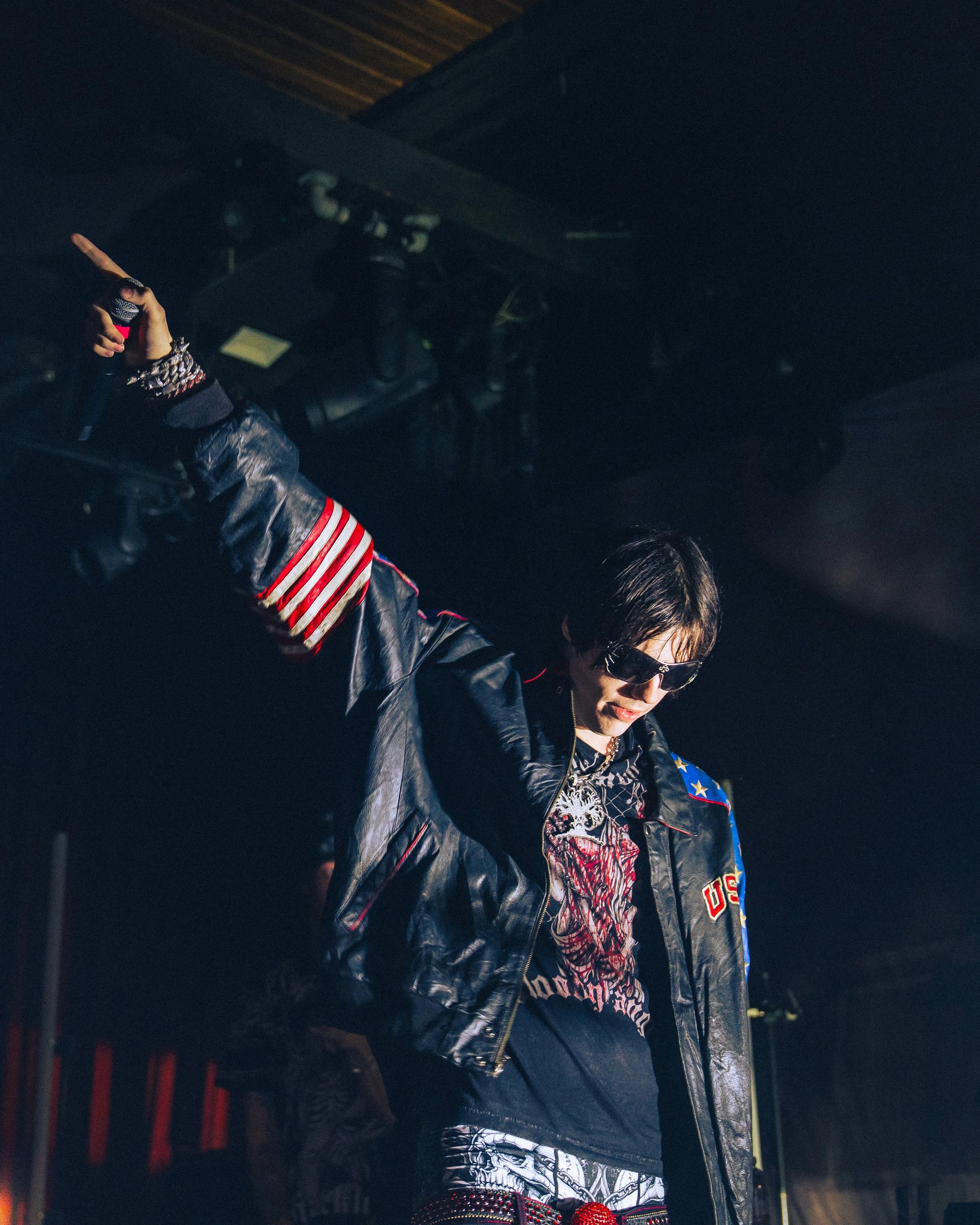 Person wearing sunglasses and a black leather jacket with military patches, holding a microphone, at night.