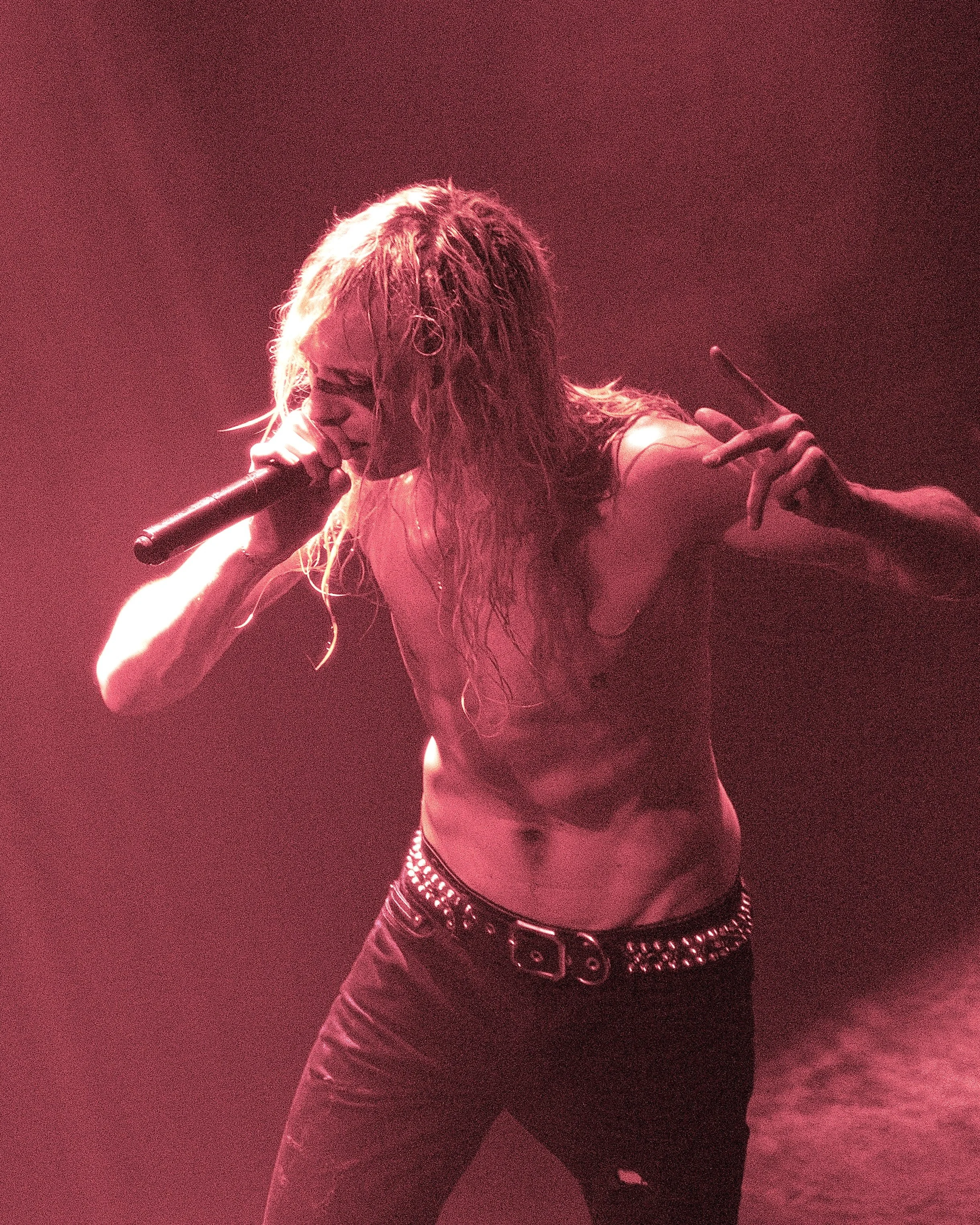 A performer with long blonde hair is singing into a microphone. She is wearing a studded belt and dark pants, dancing on stage with red lighting creating a dramatic atmosphere.