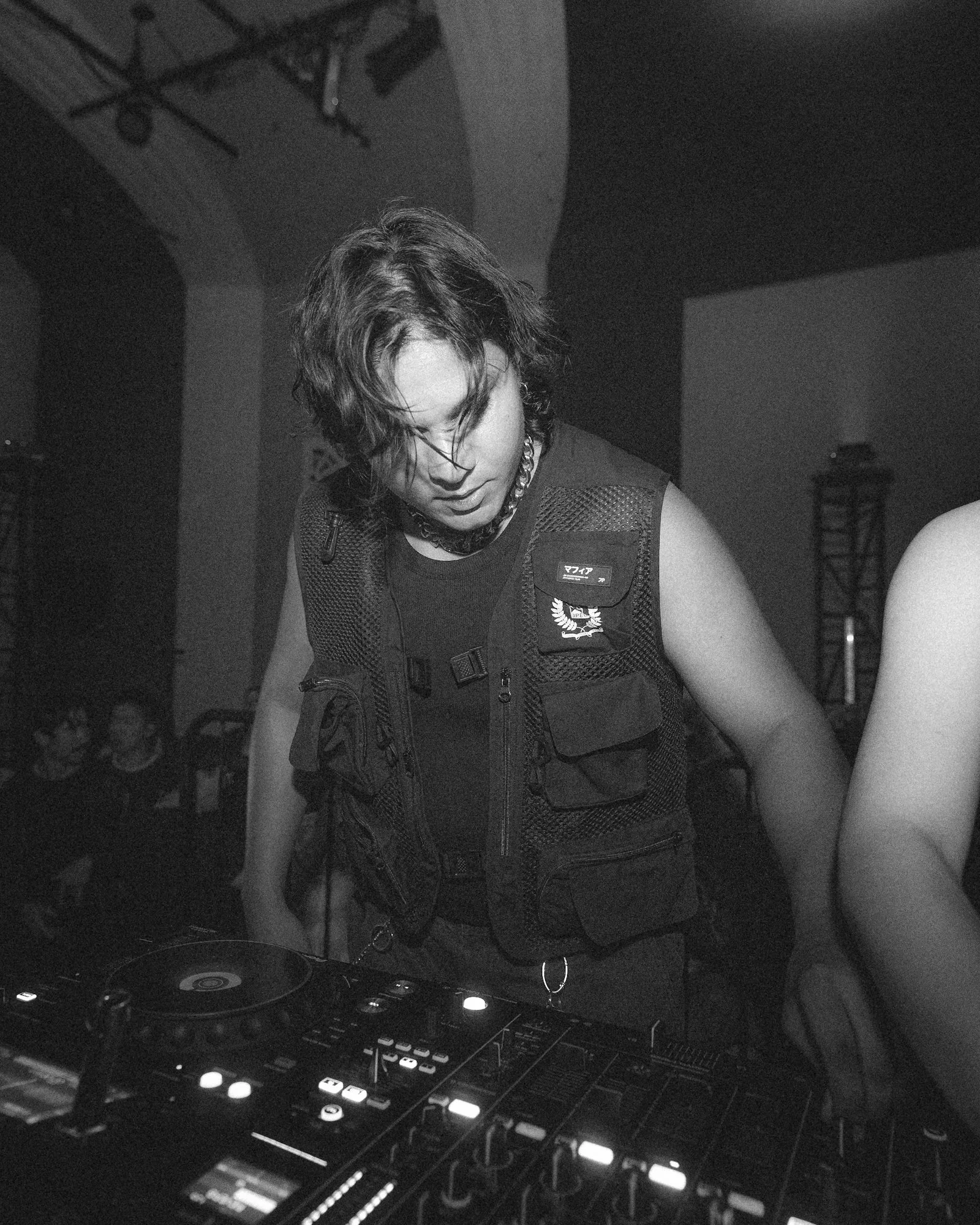 A person with wavy hair wearing a necklace and a vest, standing at a DJ mixer or turntable setup, with turntable and electronic controls, in a dimly lit room.