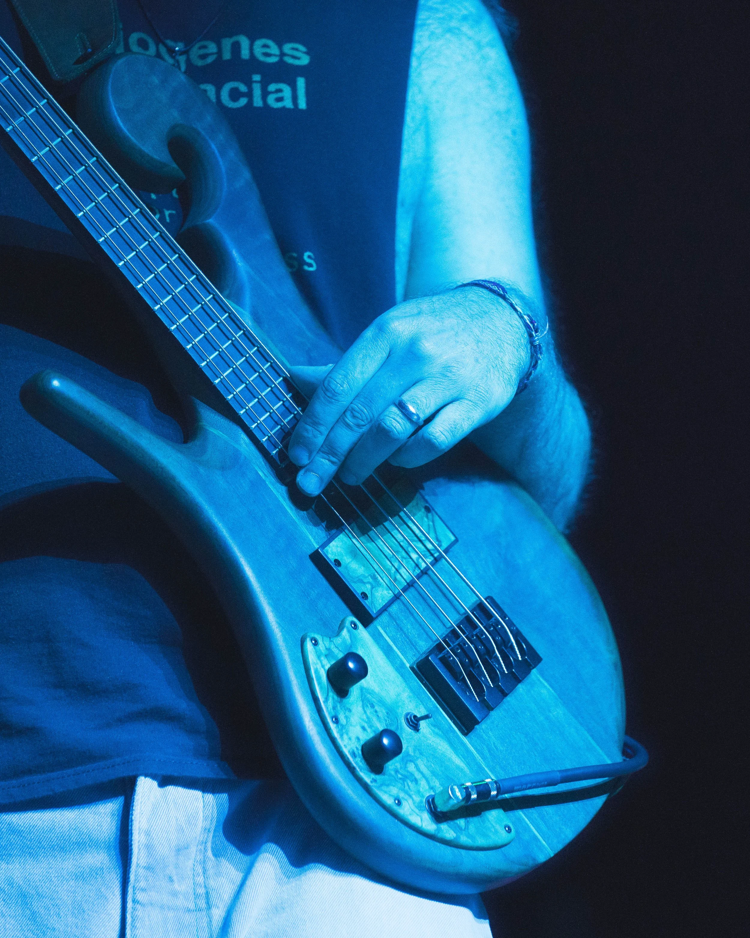 Close-up of a person playing an electric guitar, with focus on the hand on the fretboard and the guitar's body. The person is wearing a bracelet and a ring. The background is dark, and part of a shirt with partially visible text that reads 'Jovenes' 