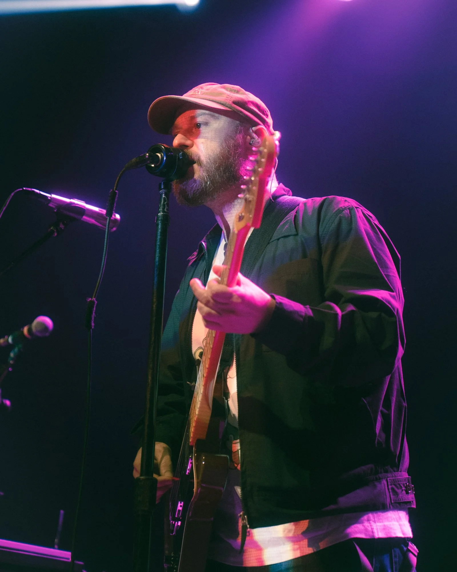 Musician with a beard and hat, singing into a microphone while playing an electric guitar on stage with purple and pink stage lighting.