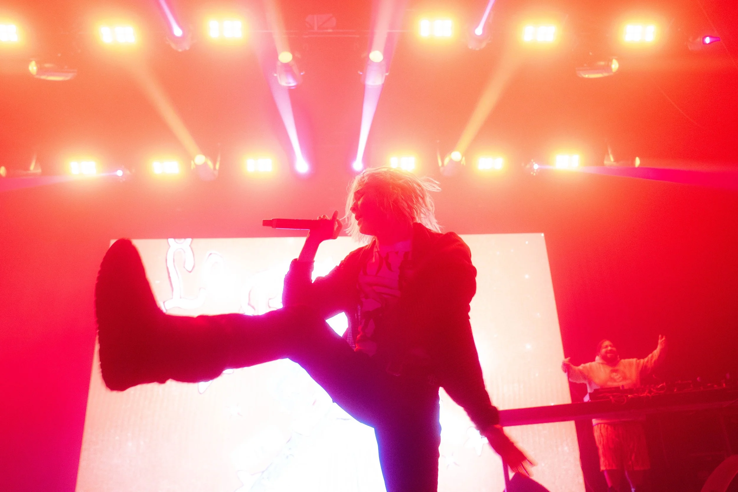Performer with messy hair singing into a microphone on stage with bright colorful lights overhead and an LED screen behind.