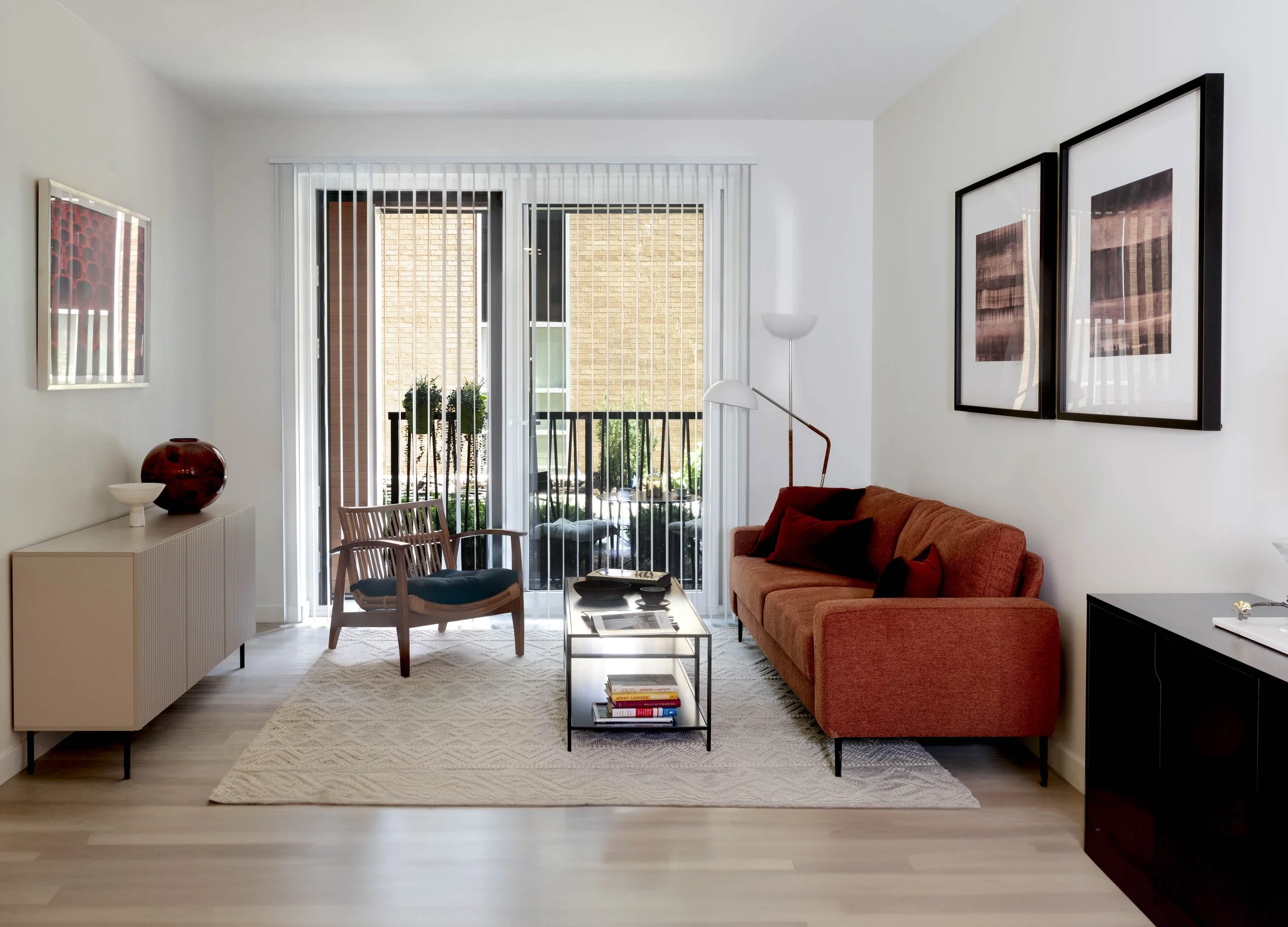 A modern living room with a sliding glass door leading to a balcony. The room has a beige cabinet with decorative bowls, a wooden chair with a dark cushion, a burnt orange sofa with black and orange pillows, a black sideboard, and wall art. There is 