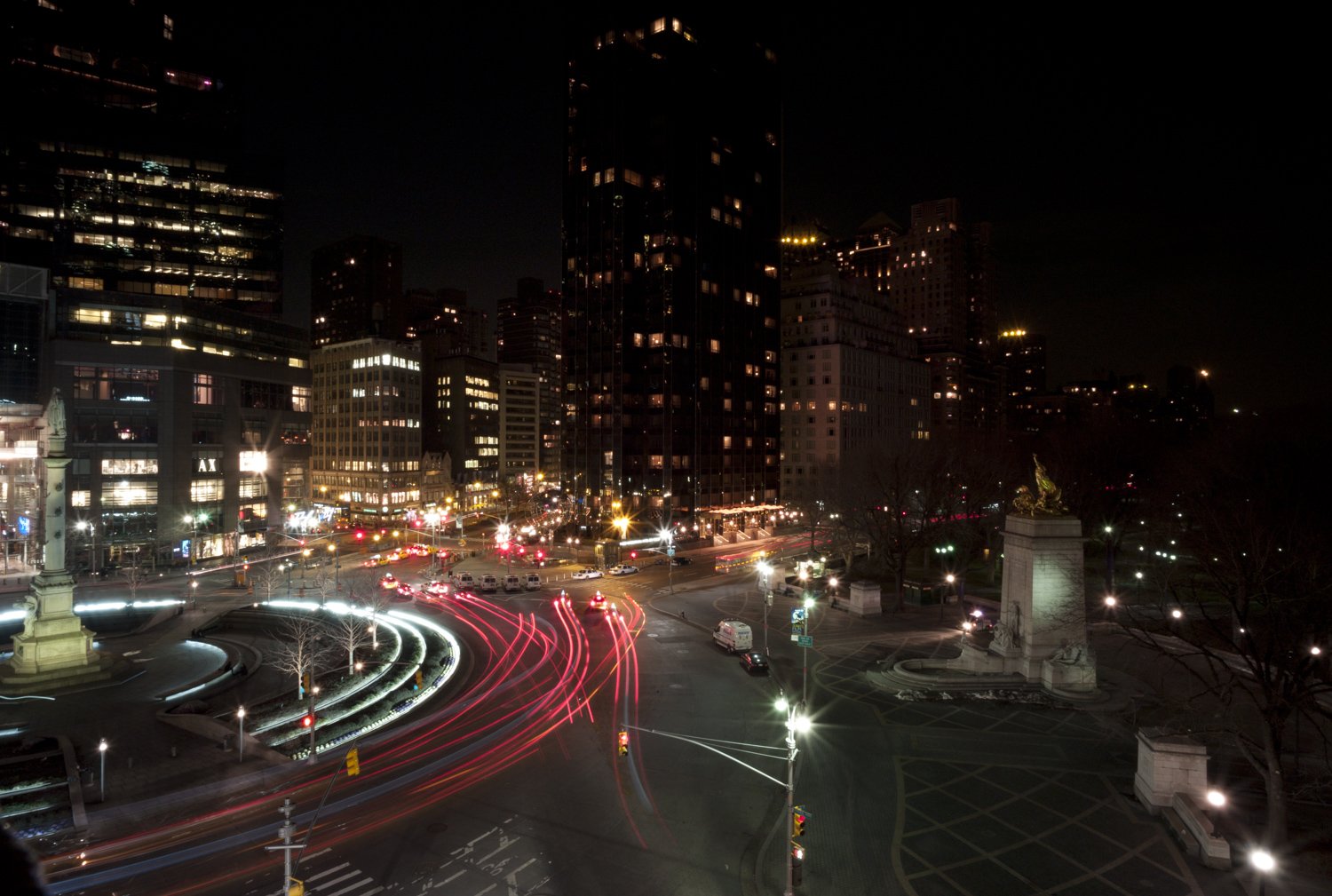 Night view of a busy city intersection with light trails from vehicles, tall buildings, and a monument in the foreground.