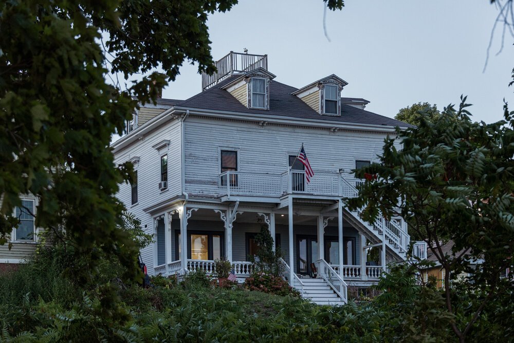 Large white colonial-style house with American flag, surrounded by trees.