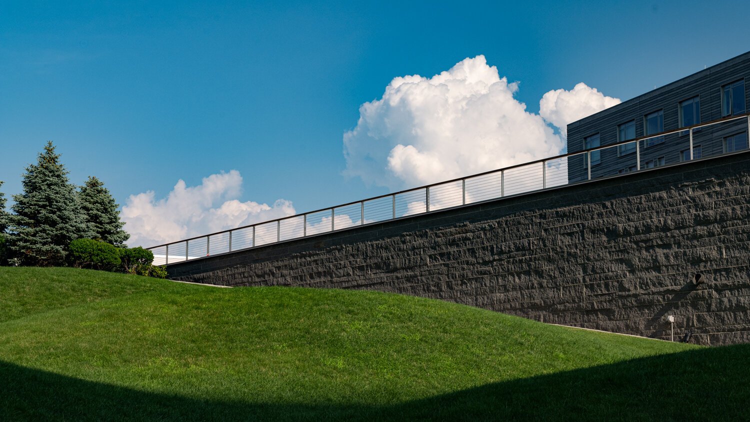 Green grassy hill with spruce trees and a building with a stone retaining wall under a blue sky with clouds.