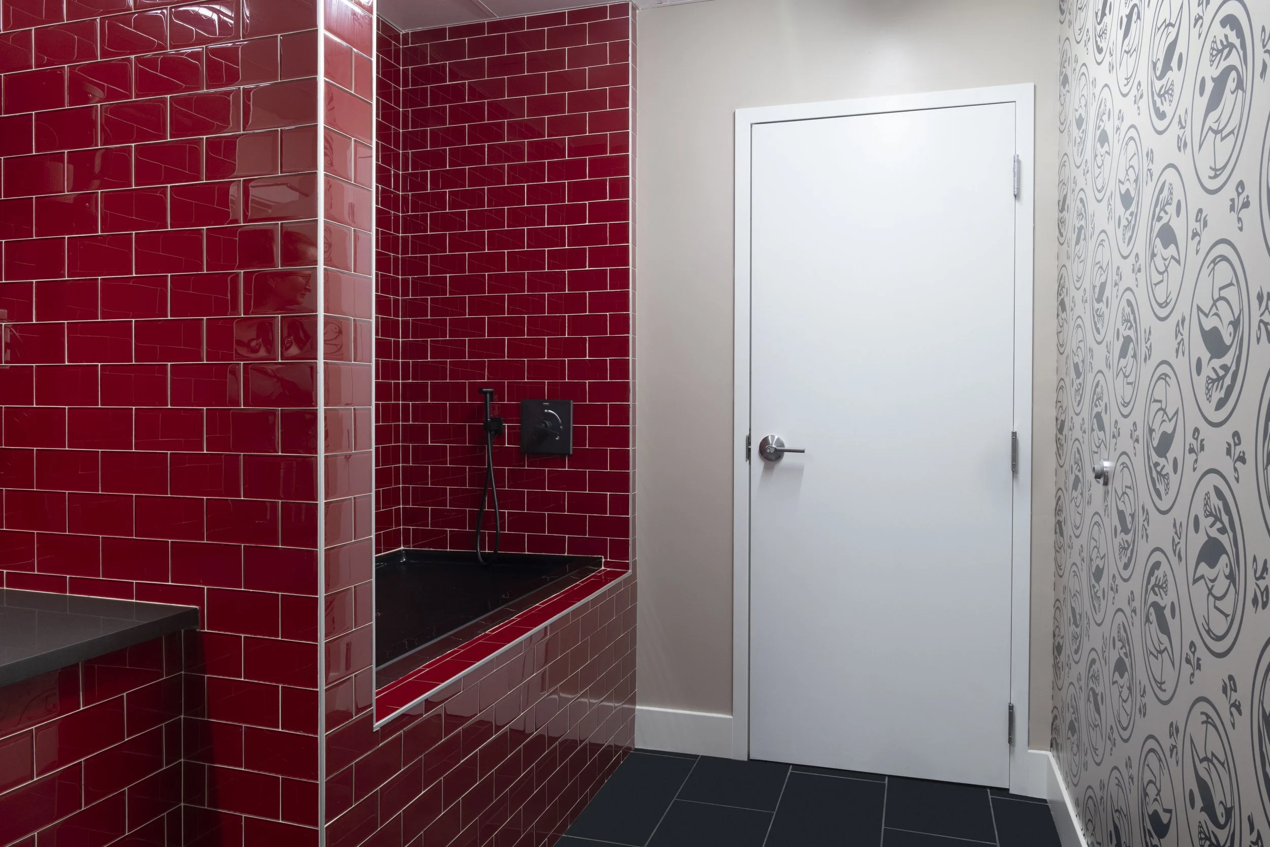 A dog wash facility with a red tiled shower area, black fixtures, and a white door. The wall on the right features patterned wallpaper with circular motifs from the property’s branding.