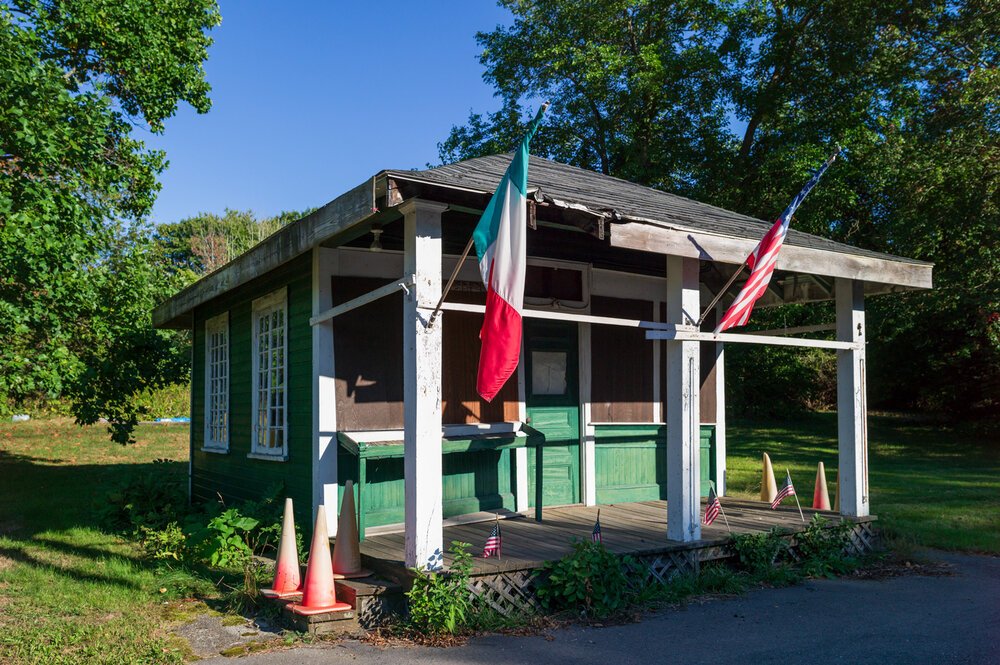 Small green wooden building with American and Italian flags, surrounded by trees and orange traffic cones.