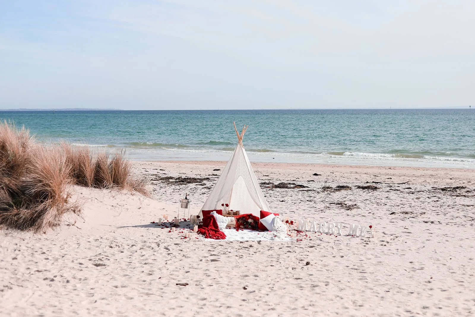 Beach scene with a small white tent decorated in red and white, surrounded by candles and pillows, with the ocean and sky in the background and sand dunes on the side.