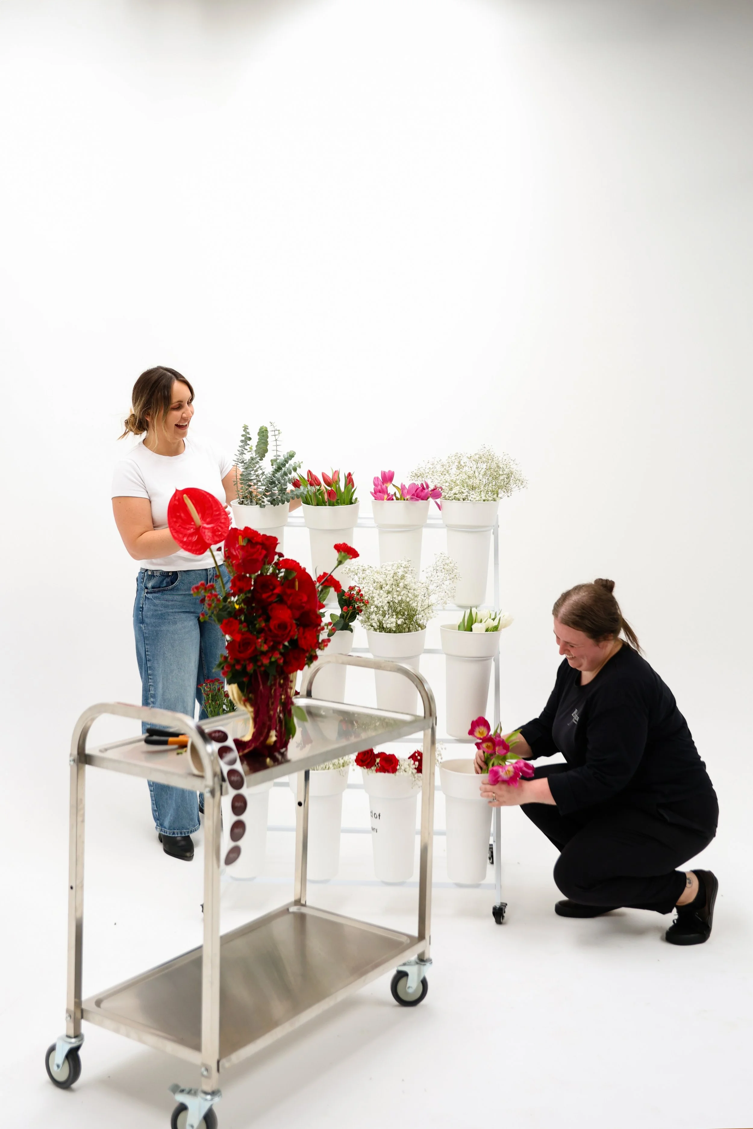 Two women arranging pink and red flowers in white pots in a white studio with minimal background.