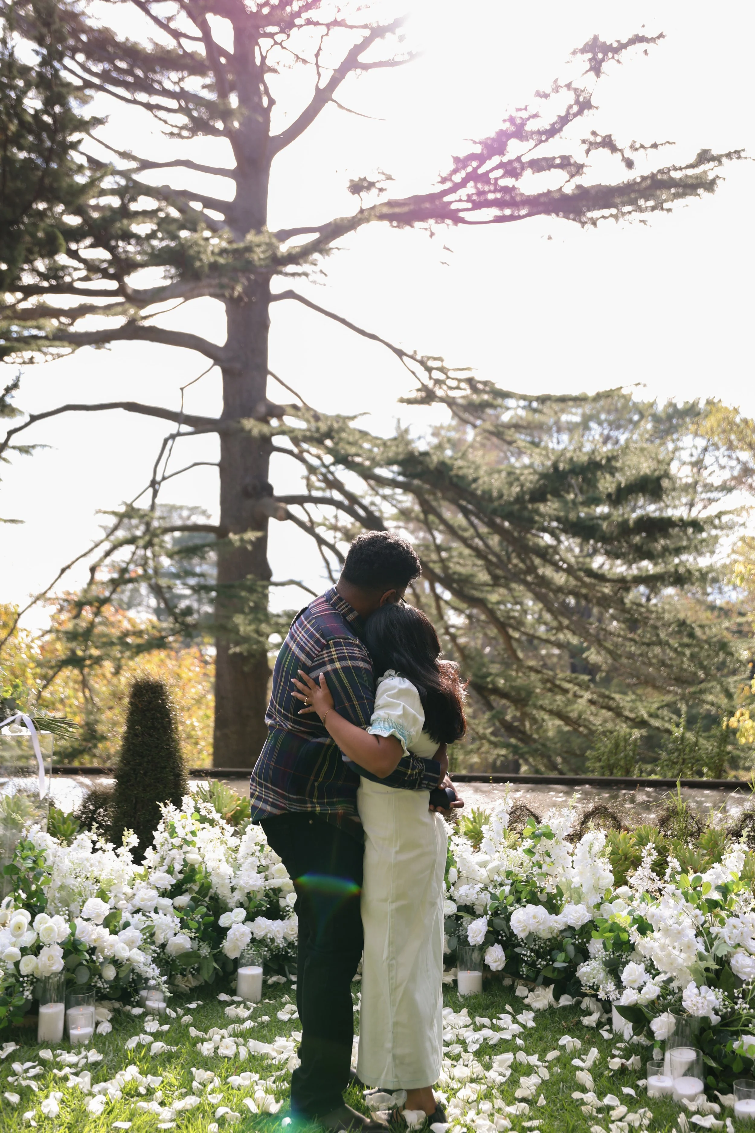 A couple is embracing at a wedding, surrounded by white flowers and candles, outdoors under trees, with sunlight streaming through the branches.