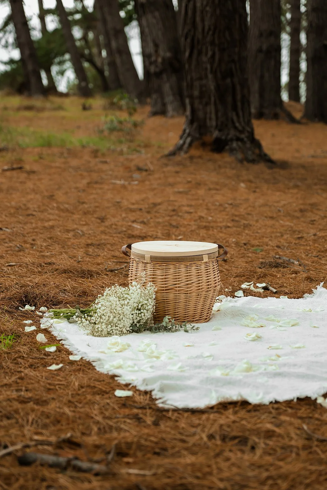 A wicker basket with a wooden lid on a white cloth with flower petals and a bouquet of white flowers, set in a wooded area.