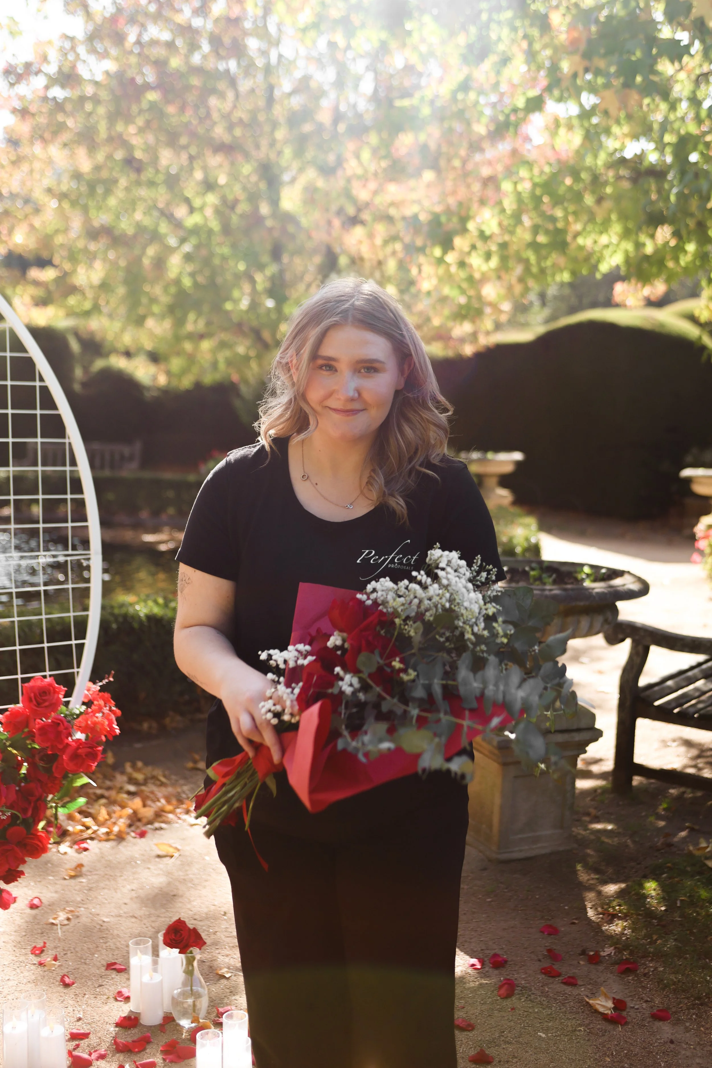 A young woman with shoulder-length blonde hair, wearing a black shirt, is holding a bouquet of red and white flowers in a red wrapping. She is outdoors in a park, with sunlight filtering through trees with green and yellow leaves. There are white candles on the ground surrounded by rose petals near her.