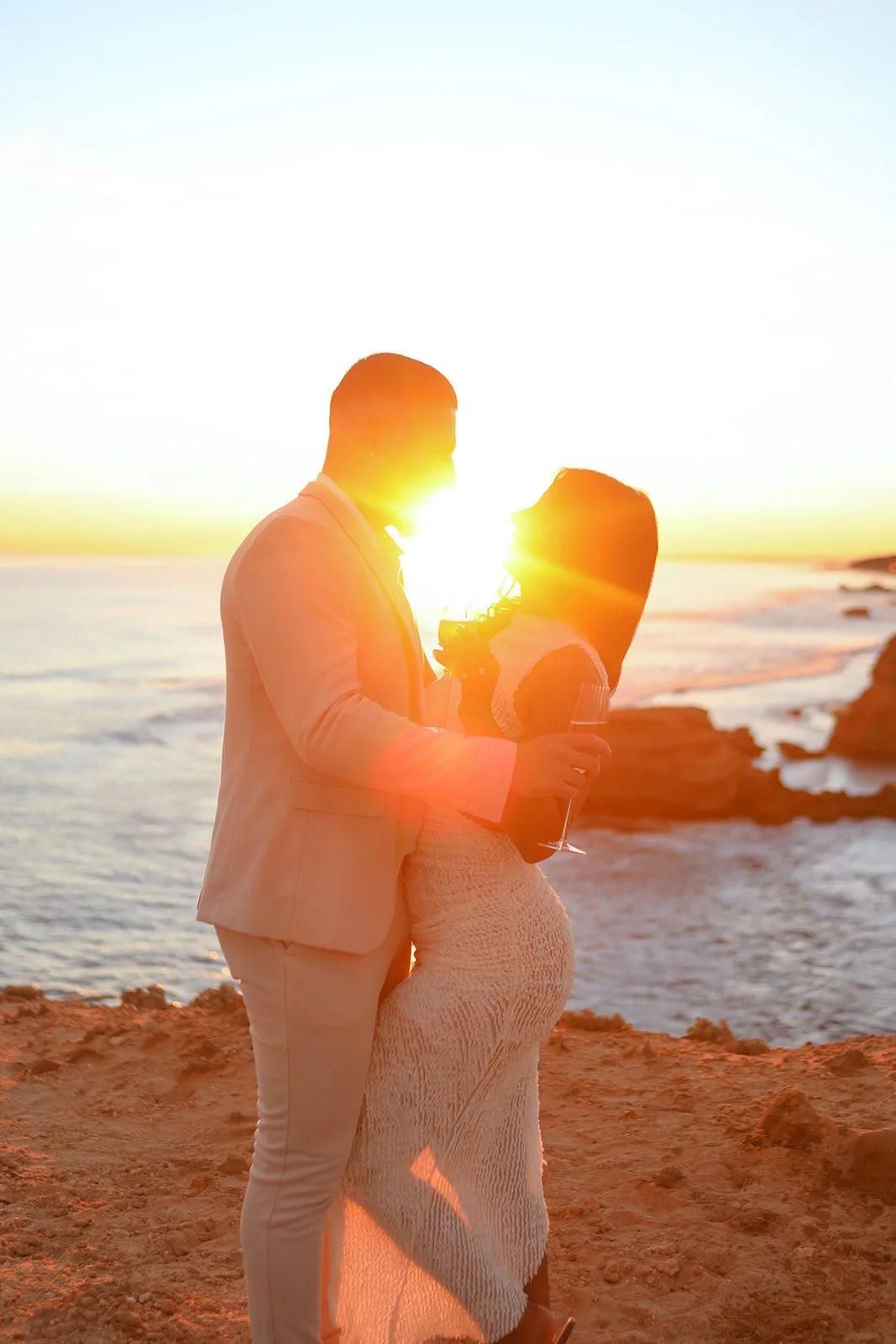 A couple dressed in formal attire standing on a beach at sunset, looking at each other with the sun setting behind them.