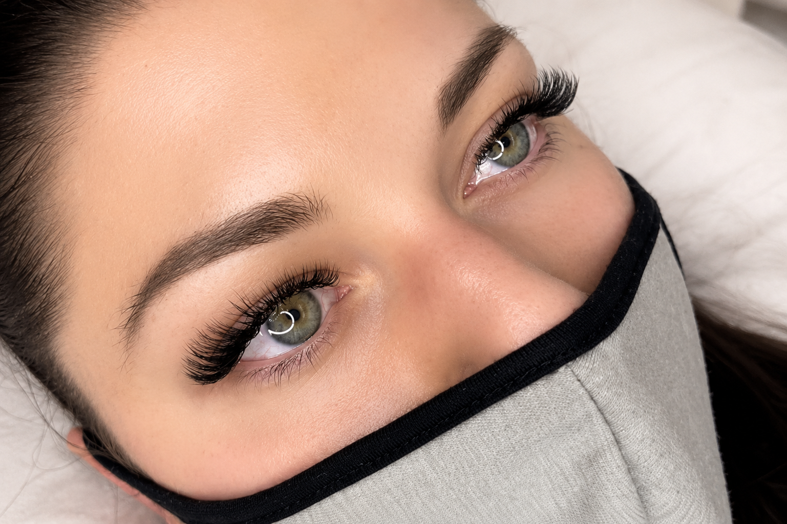 Close-up of a woman with hazel eyes, makeup, and long lashes wearing a black face mask, lying down on a pillow at Tenderoni Lashes located in Los Angeles.