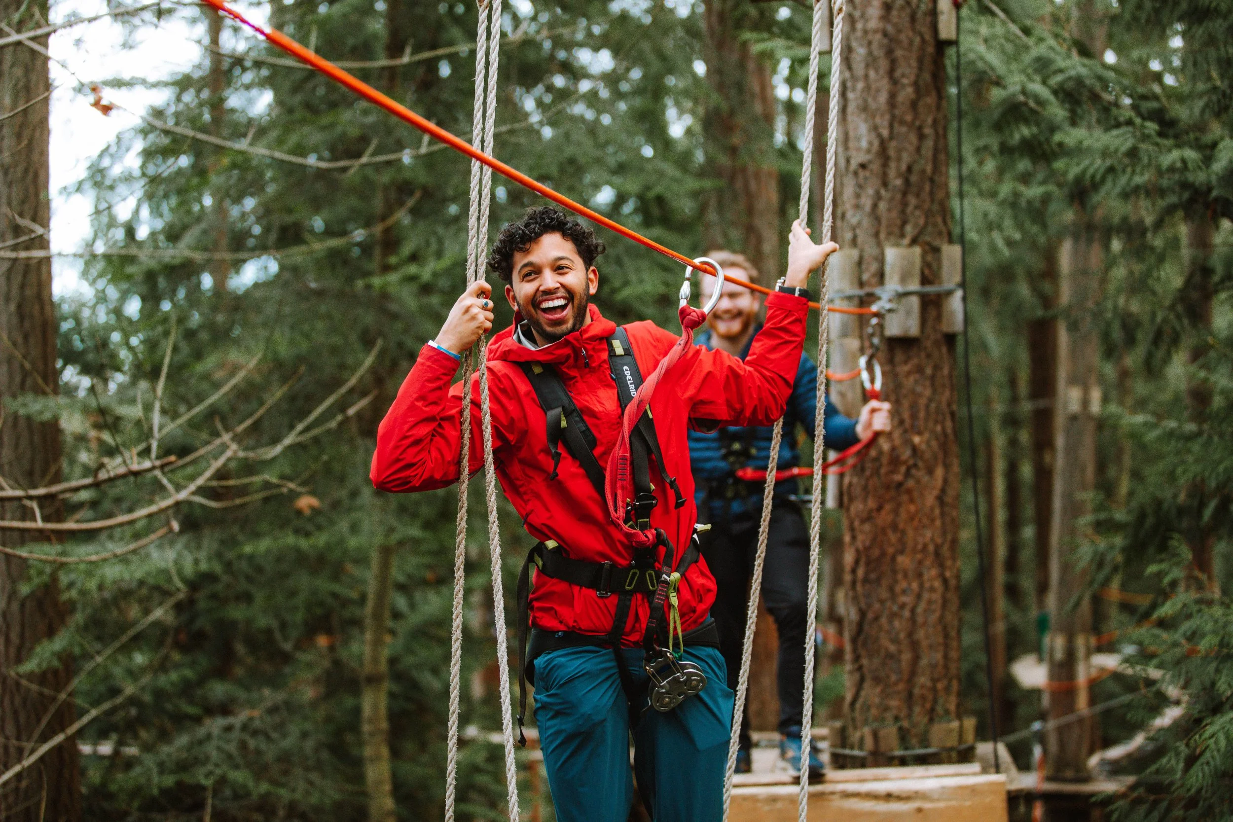 Two men in a forest on ropes and wooden platforms, enjoying a canopy adventure course. The man in front is smiling and holding onto a rope, wearing a red jacket, blue pants, and safety harness.